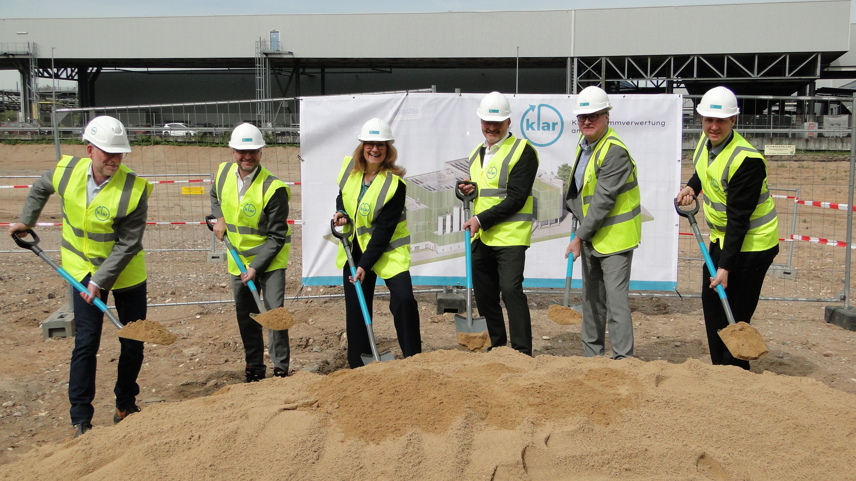Zu sehen sind sechs Personen, die Warnwesten und Schutzhelme tragen, und gemeinsam einen ersten Spatenstich machen.