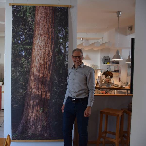 Ein Mann steht in einem Café, neben einem großformatigen Bild eines Baumstammes.