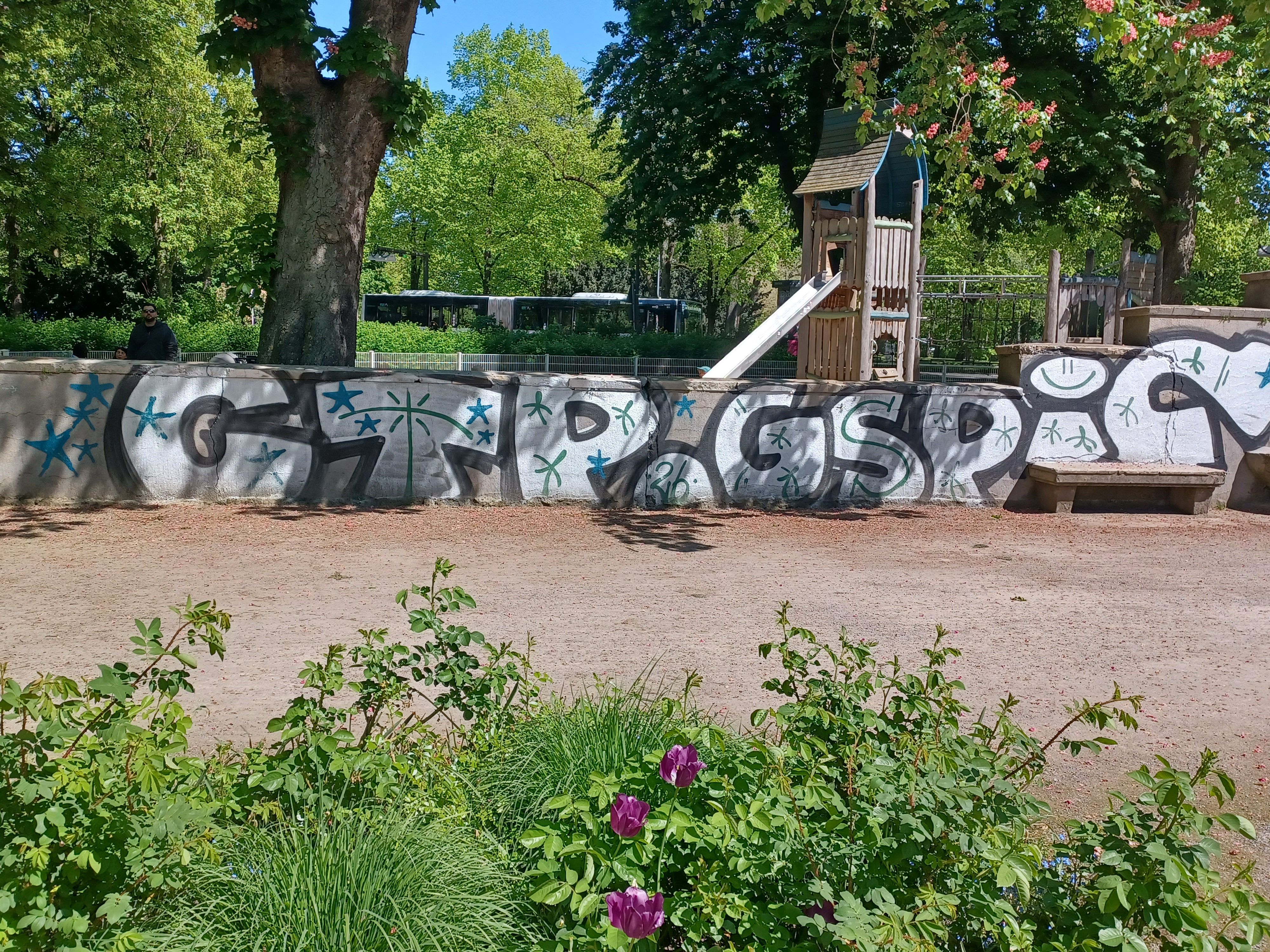 Blick auf eine mit Graffiti besprühte Mauer, dahinter liegt ein Spielplatz.