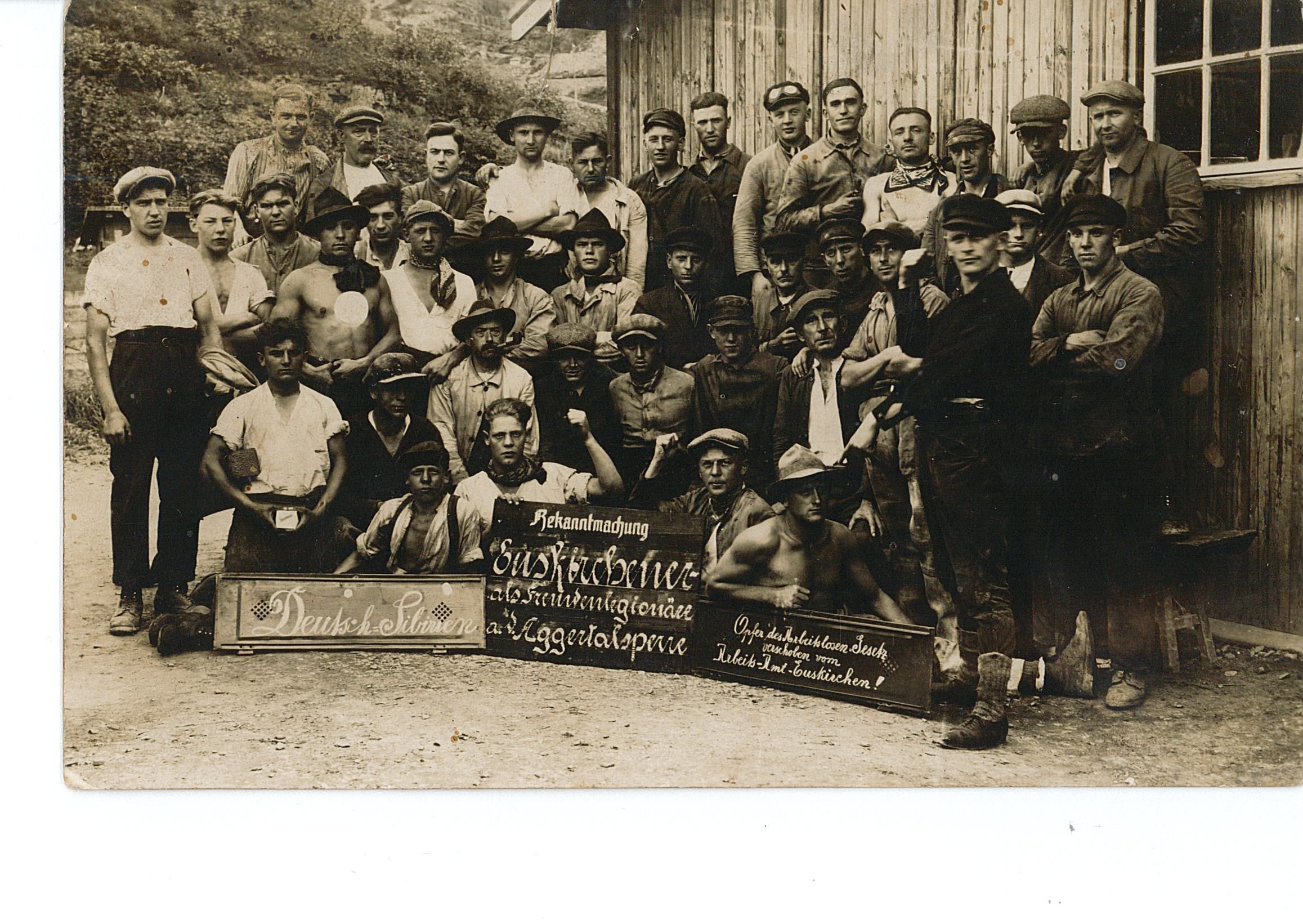 Dies ist die Vorderseite einer nicht gelaufenen Postkarte. Die Fotografie zeigt offensichtlich unzufriedene Arbeiter aus dem Raum Euskirchen, die sich laut einem Schild als „Opfer des Arbeitslosengesetzes“ sehen, beim Bau der Aggertalsperre bei Gummersbach, vermutlich 1927.