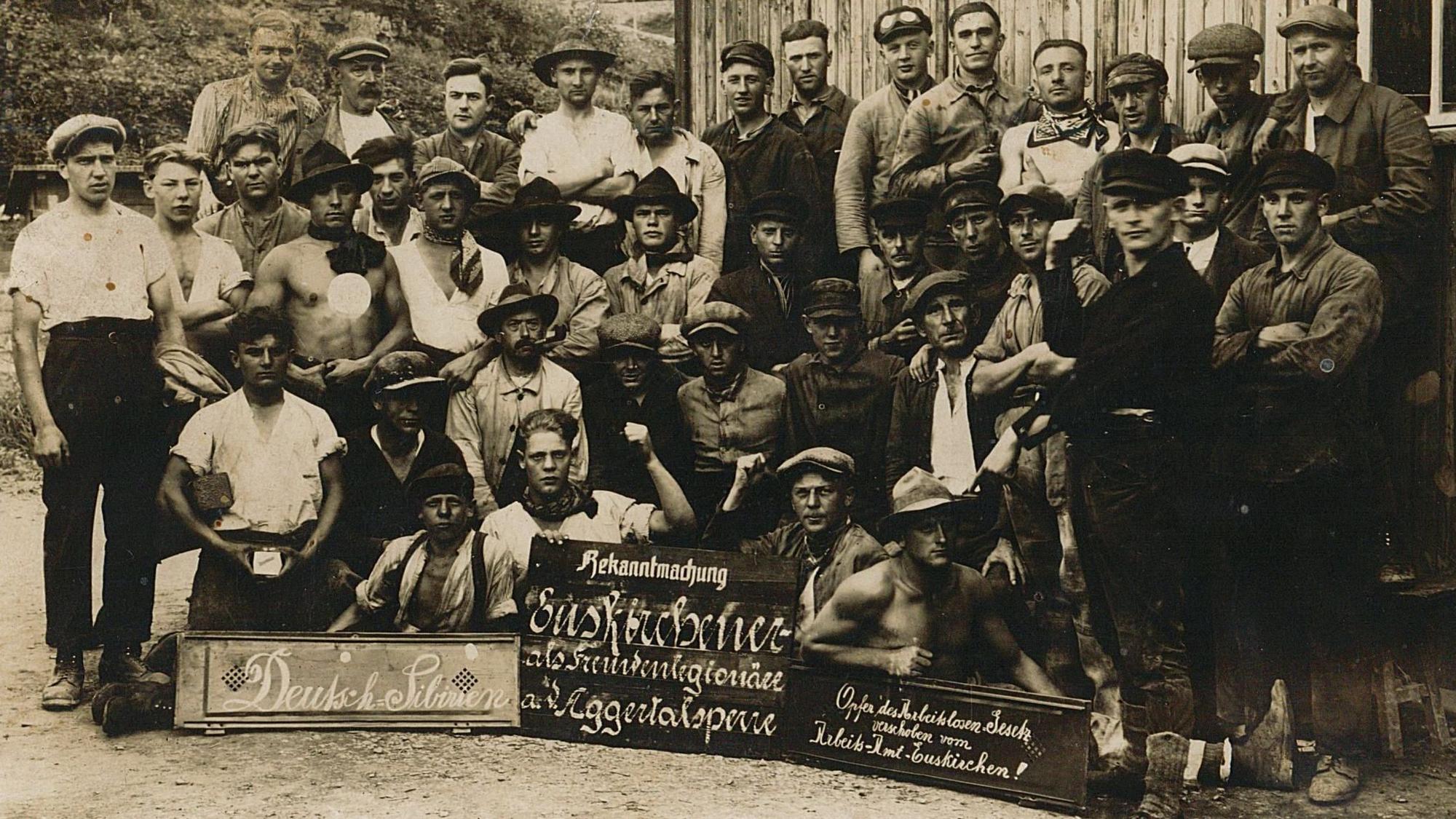 Dies ist die Vorderseite einer nicht gelaufenen Postkarte. Die Fotografie zeigt offensichtlich unzufriedene Arbeiter aus dem Raum Euskirchen, die sich laut einem Schild als „Opfer des Arbeitslosengesetzes“ sehen, beim Bau der Aggertalsperre bei Gummersbach, vermutlich 1927.