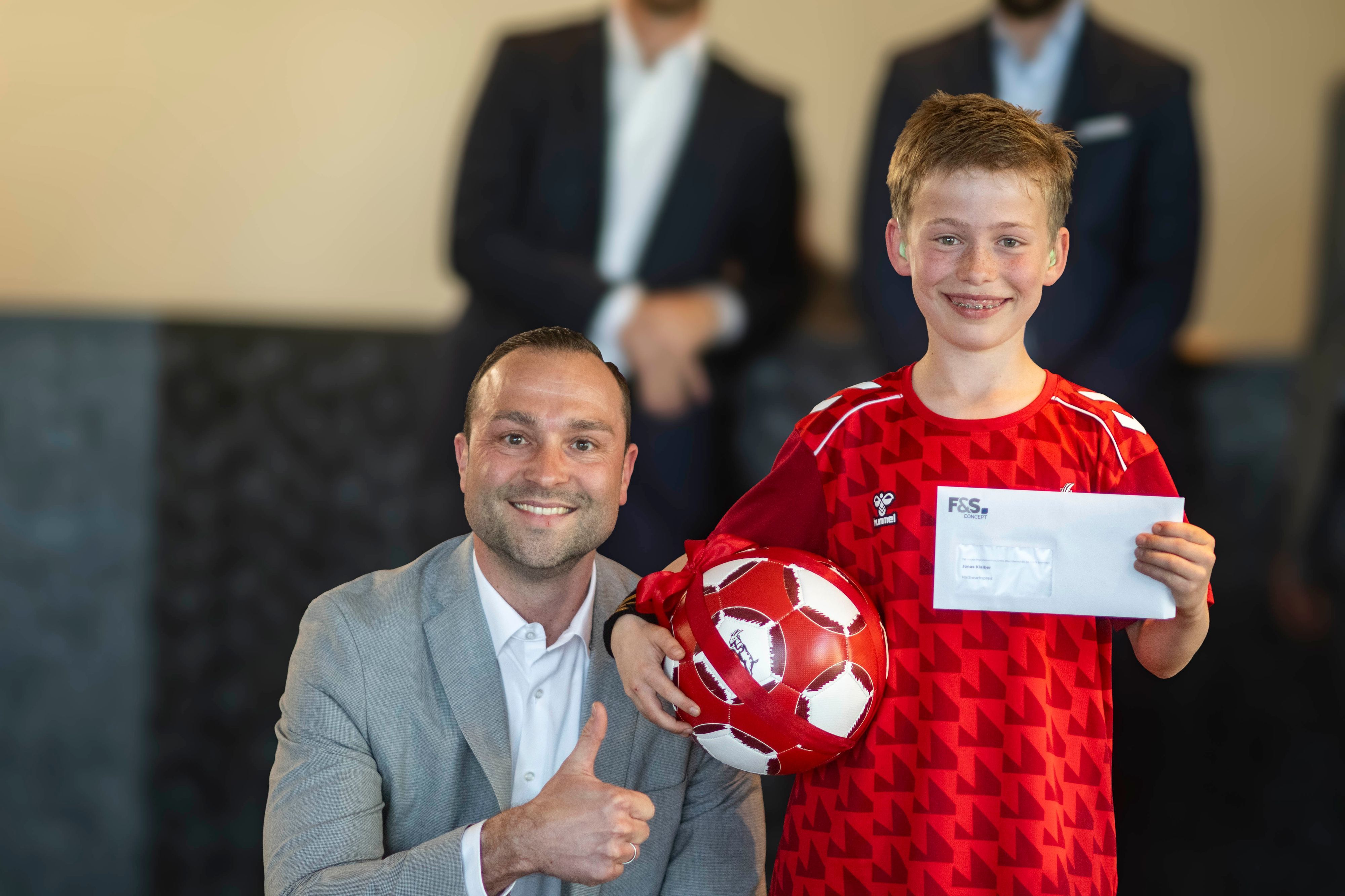 Jan Radermacher kniet neben Jonas Klaiber, der einen Fußball unterm Arm und einen Umschlag in der Hand hält.