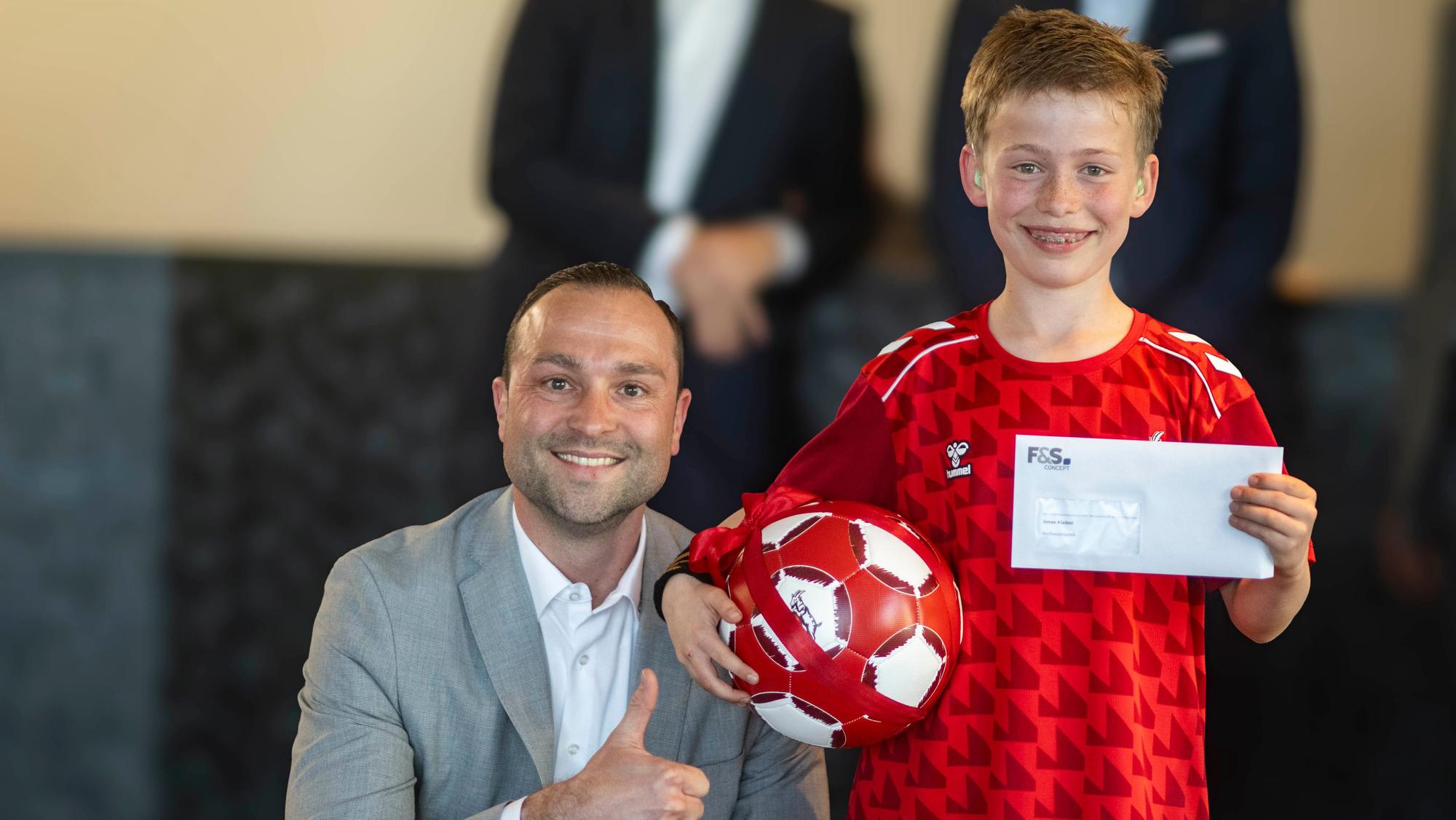 Jan Radermacher kniet neben Jonas Klaiber, der einen Fußball unterm Arm und einen Umschlag in der Hand hält.
