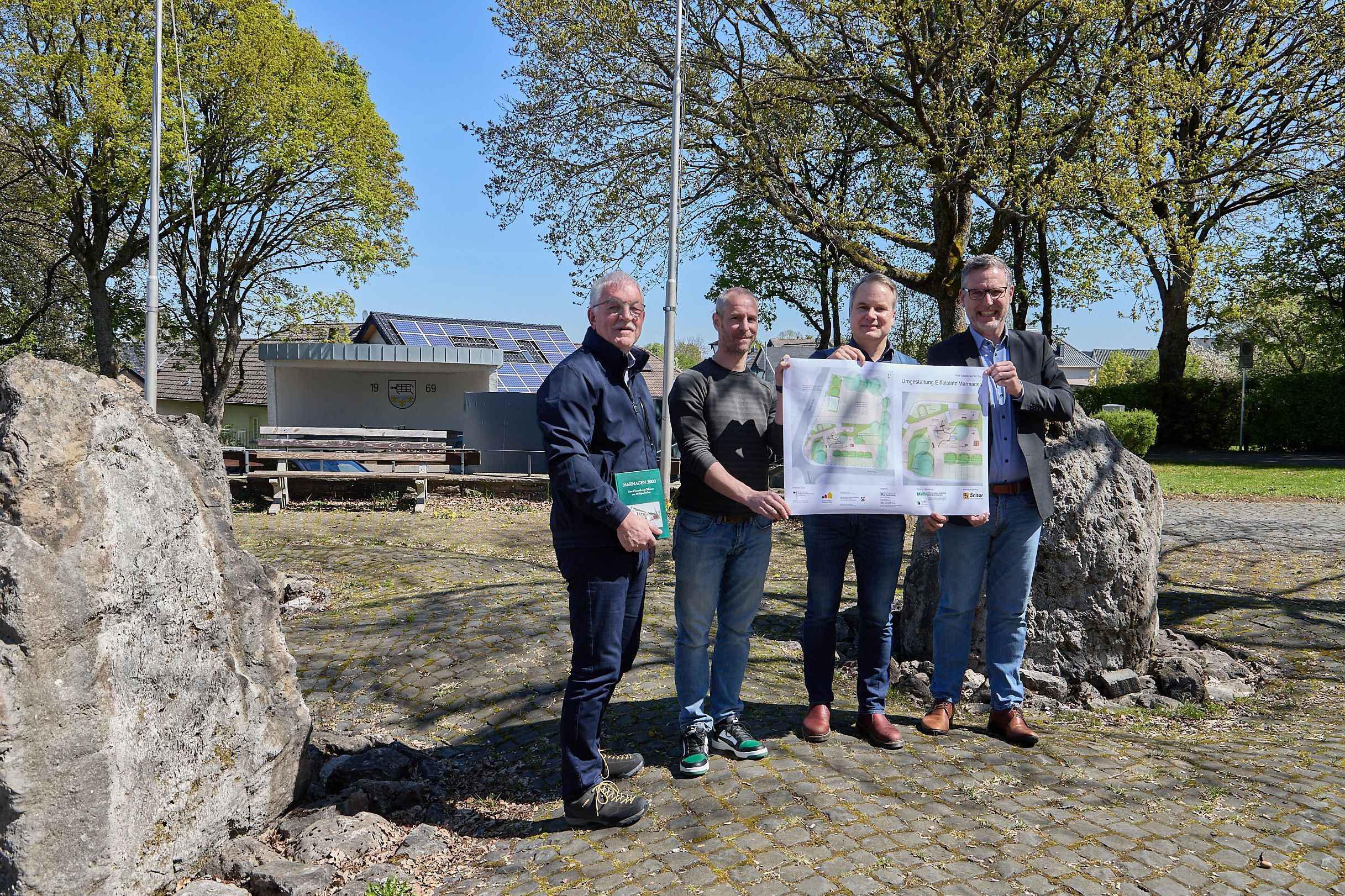Vier Männer stehen auf dem Eiffelplatz in Nettersheim und halten gemeinsam eine große Planungsskizze.