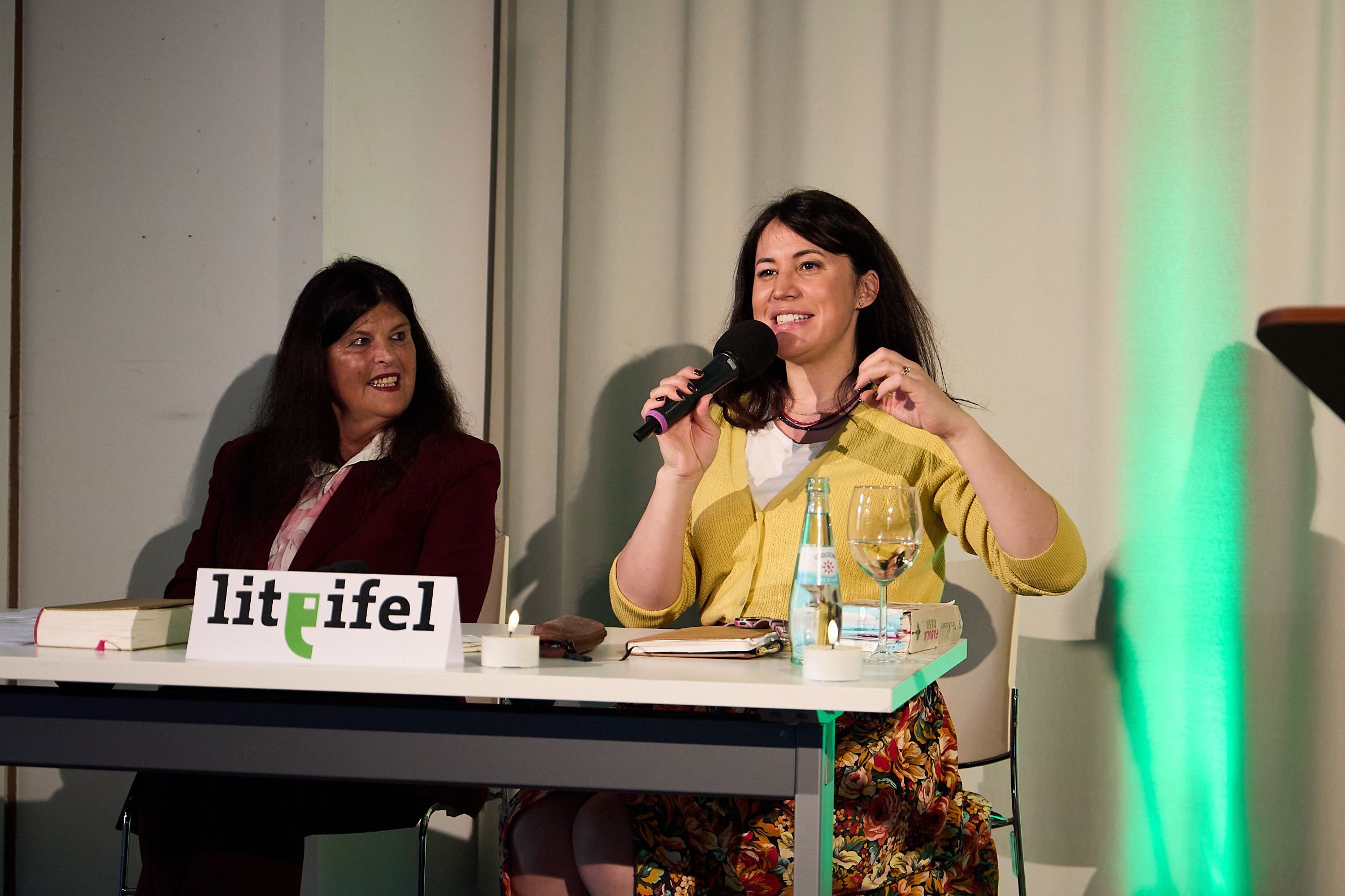 Claudia Hoffmann und Vea Kaiser sitzen an einem Tisch, auf dem ein Schild mit der Aufschrift Lit.Eifel steht. Kaiser trägt eine gelbe Jacke und gestikuliert.