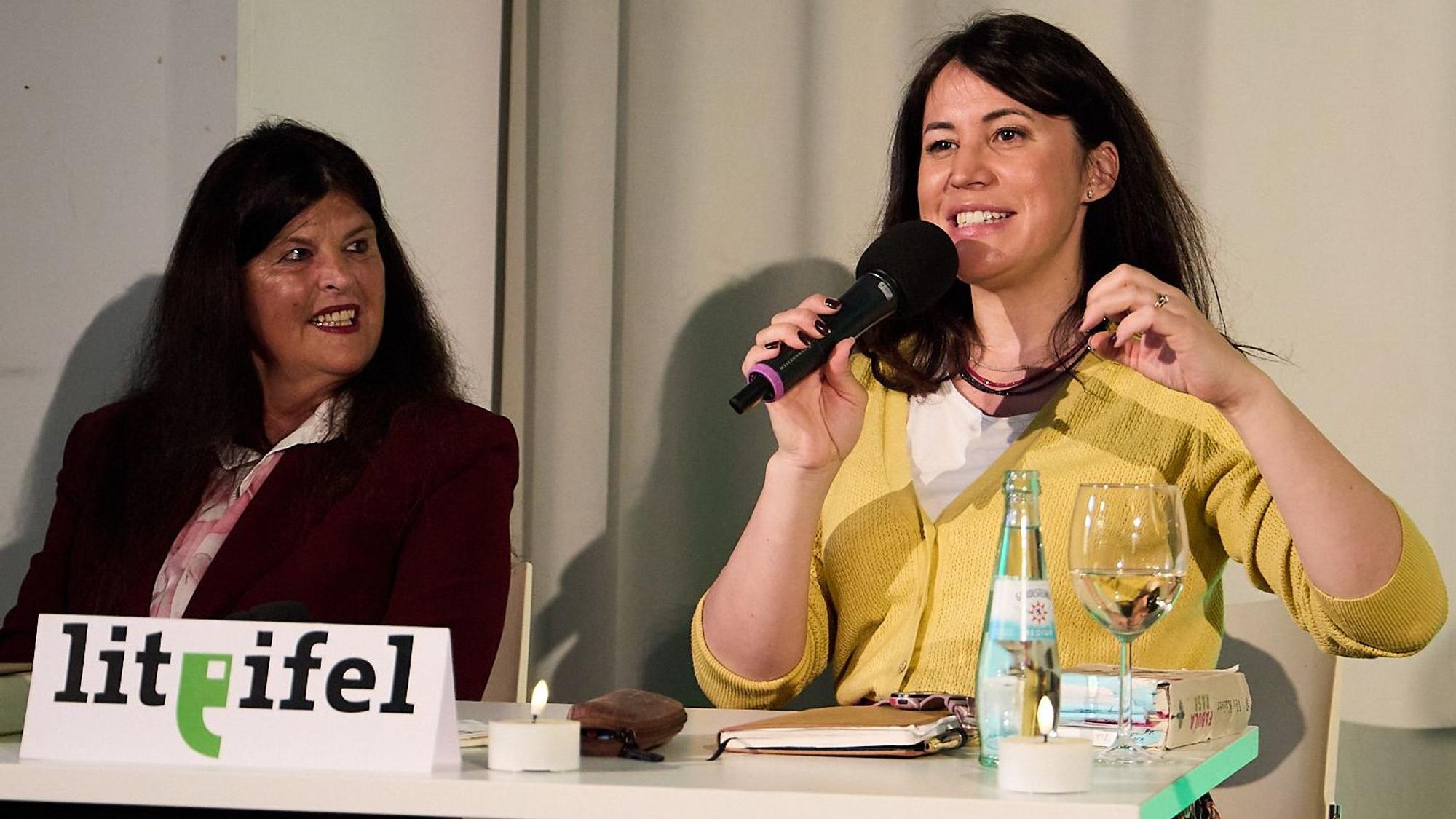 Claudia Hoffmann und Vea Kaiser sitzen an einem Tisch, auf dem ein Schild mit der Aufschrift Lit.Eifel steht. Kaiser trägt eine gelbe Jacke und gestikuliert.