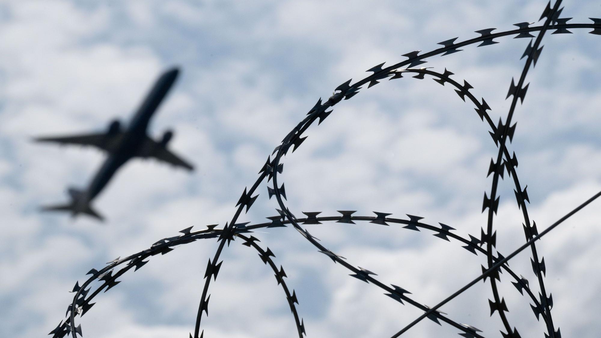 Ein Flugzeug am Himmel. Im Vordergrund ist Stacheldrahtzaun zu sehen.