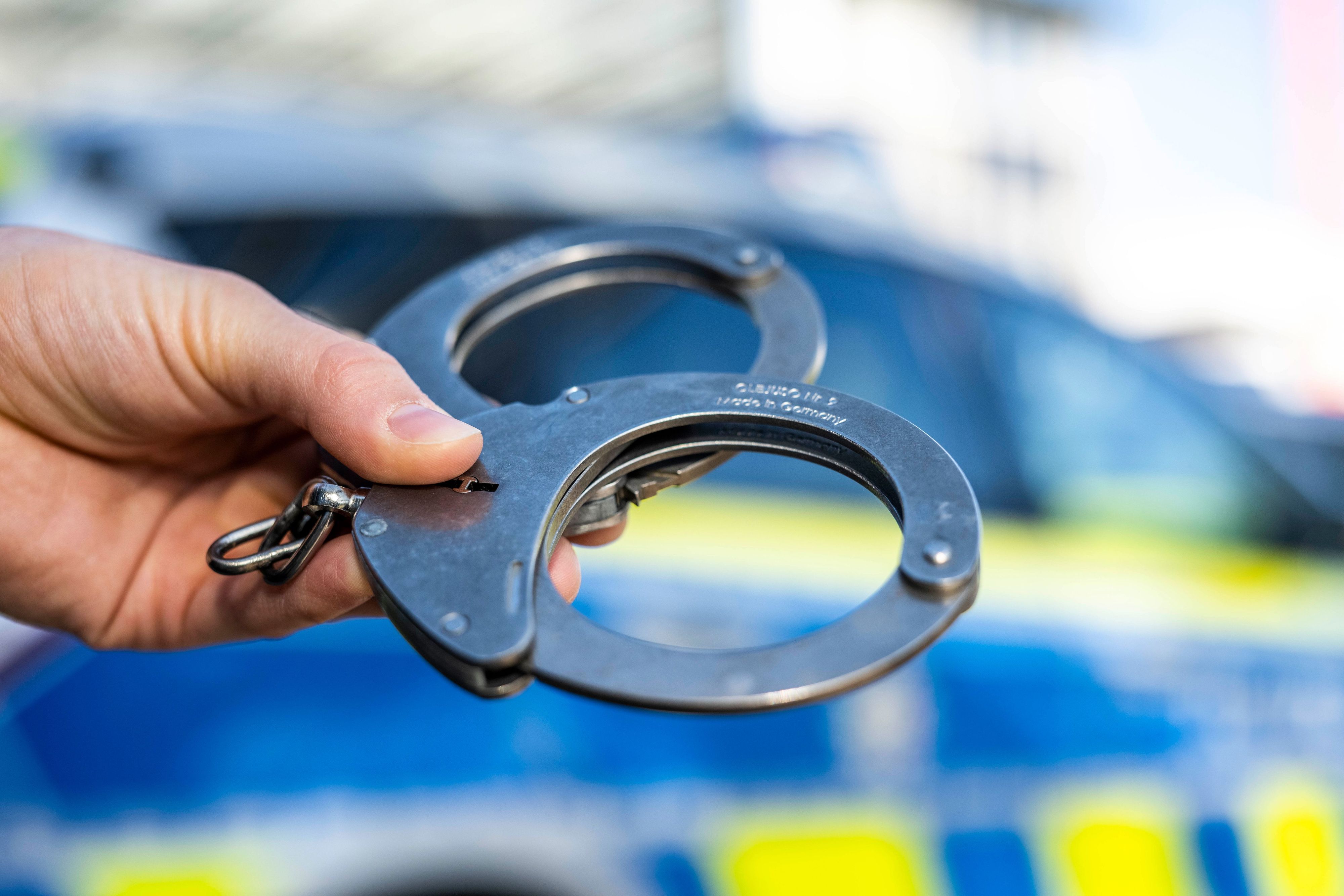 Eine Hand hält Handschellen vor einen Streifenwagen der Polizei. (Symbolbild)