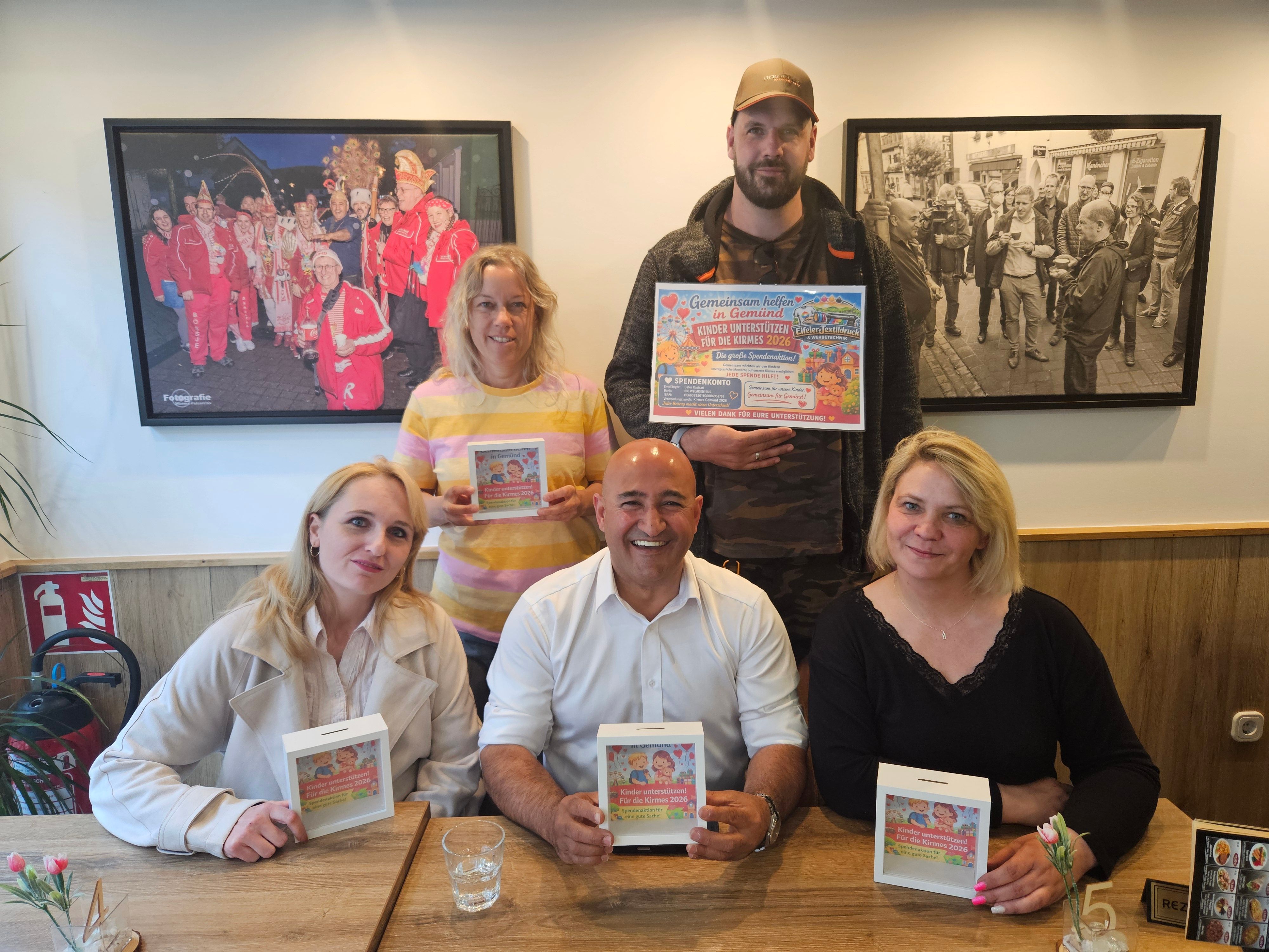 Eine Frau und ein Mann stehen hinter zwei Frauen und einem Mann, die an einem Tisch sitzen. Alle bis auf den stehenden Mann halten Spendenboxen in ihren Händen. Er hält ein Plakat hoch.