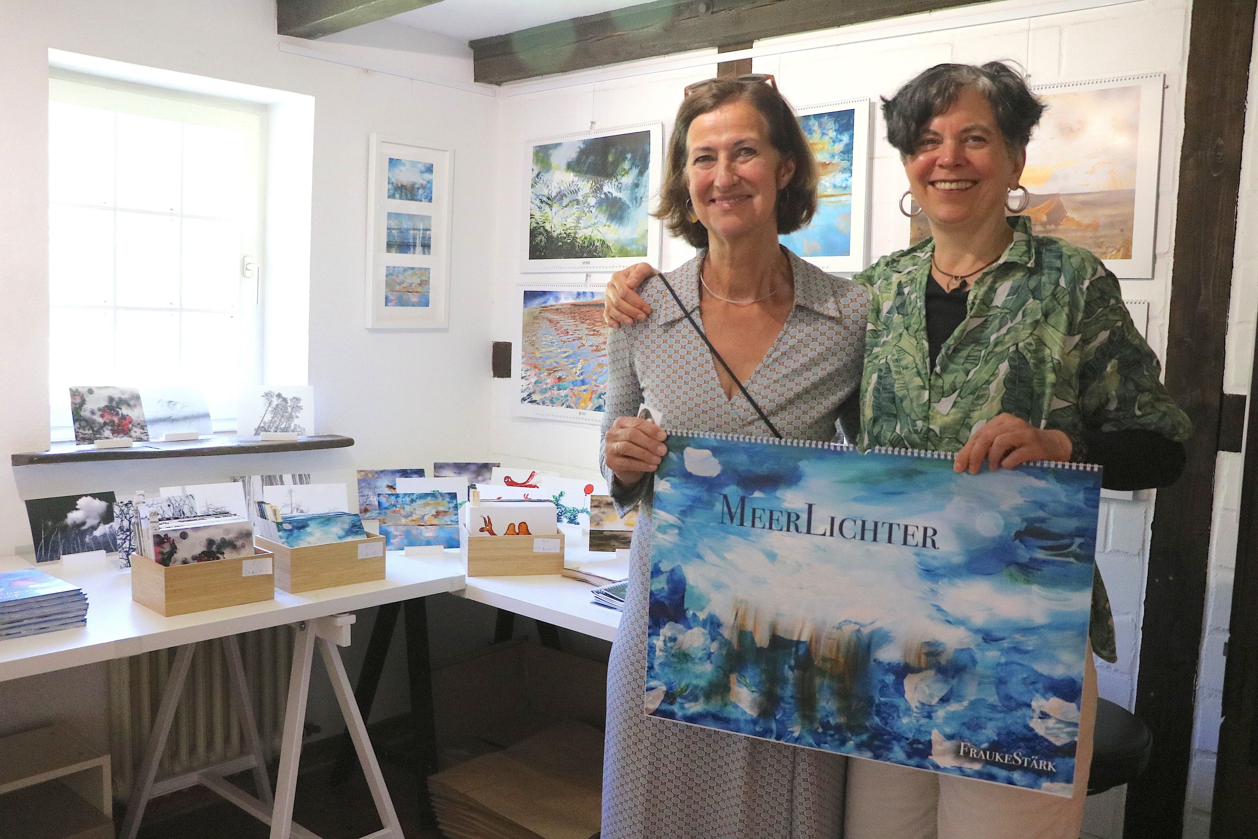 Zwei Frauen stehen nebeneinander, sie halten einen blauen Wandkalender in den Händen.
