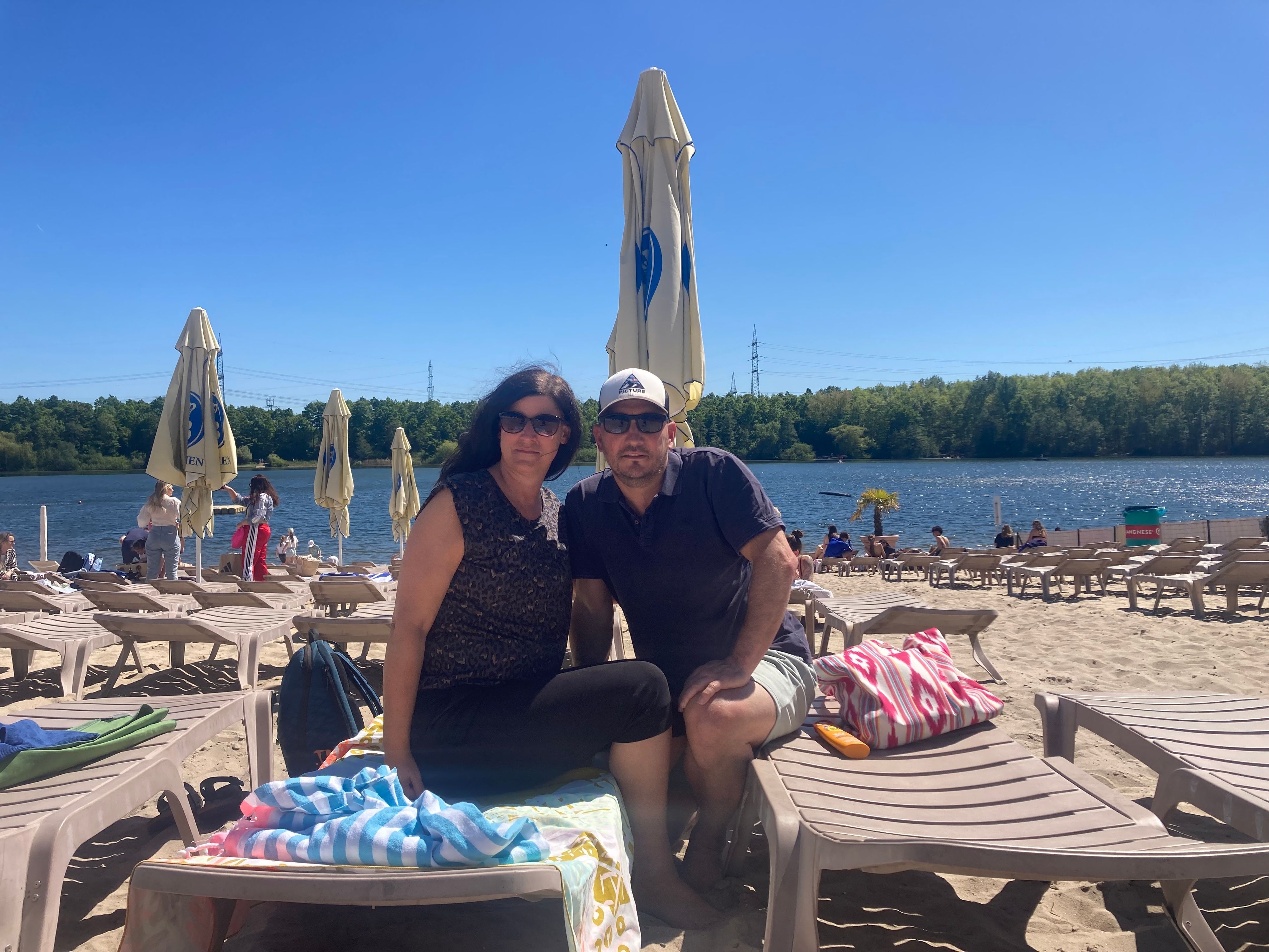 Eine Frau und ein Mann sitzen auf Strandliegen, im Hintergrund ein See.