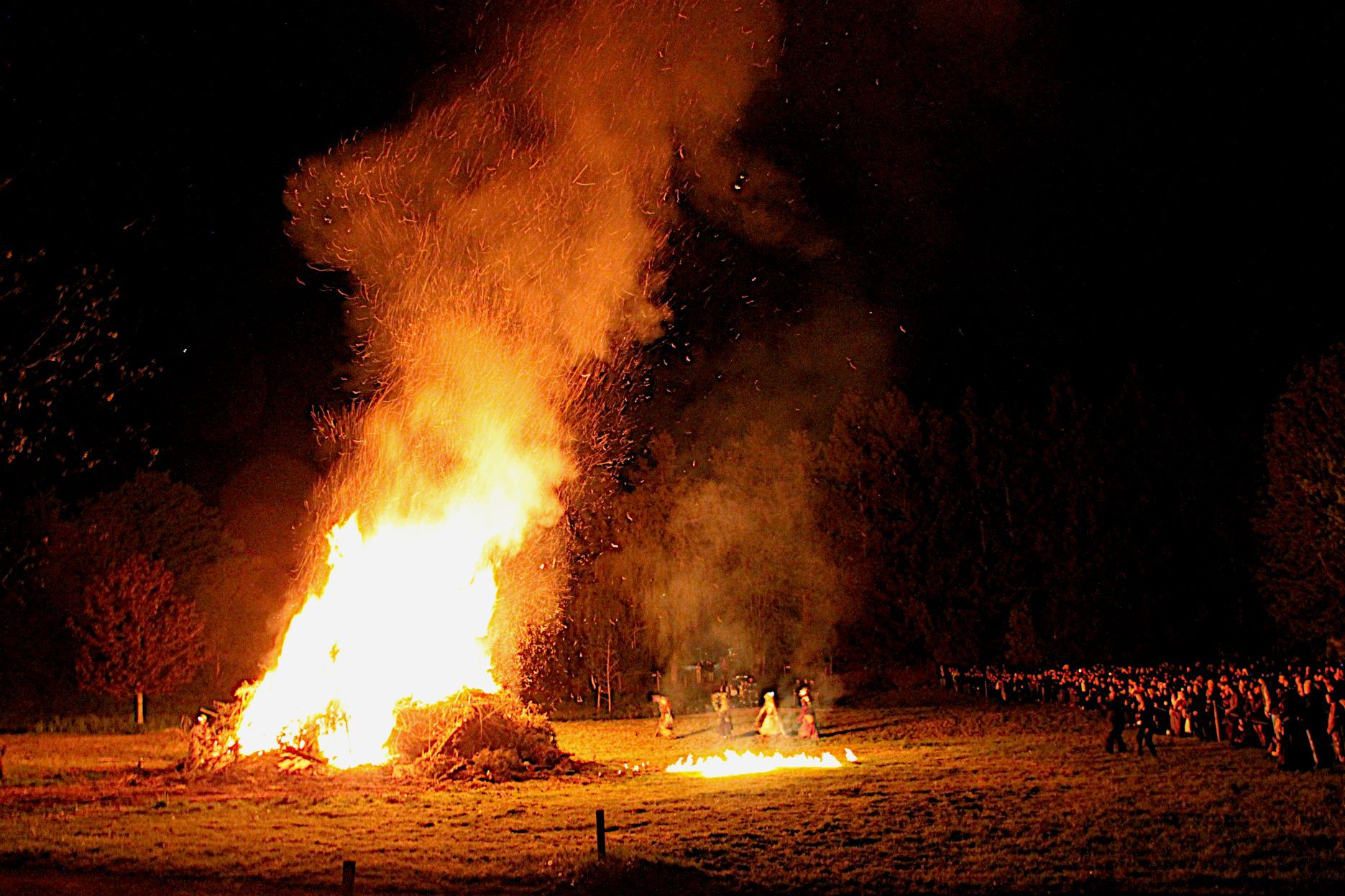 Hoch lodern die Flammen in den nächtlichen Himmel, davor tanzen die vier Hexen. An der Seite verfolgen die Zuschauer das Spektakel.