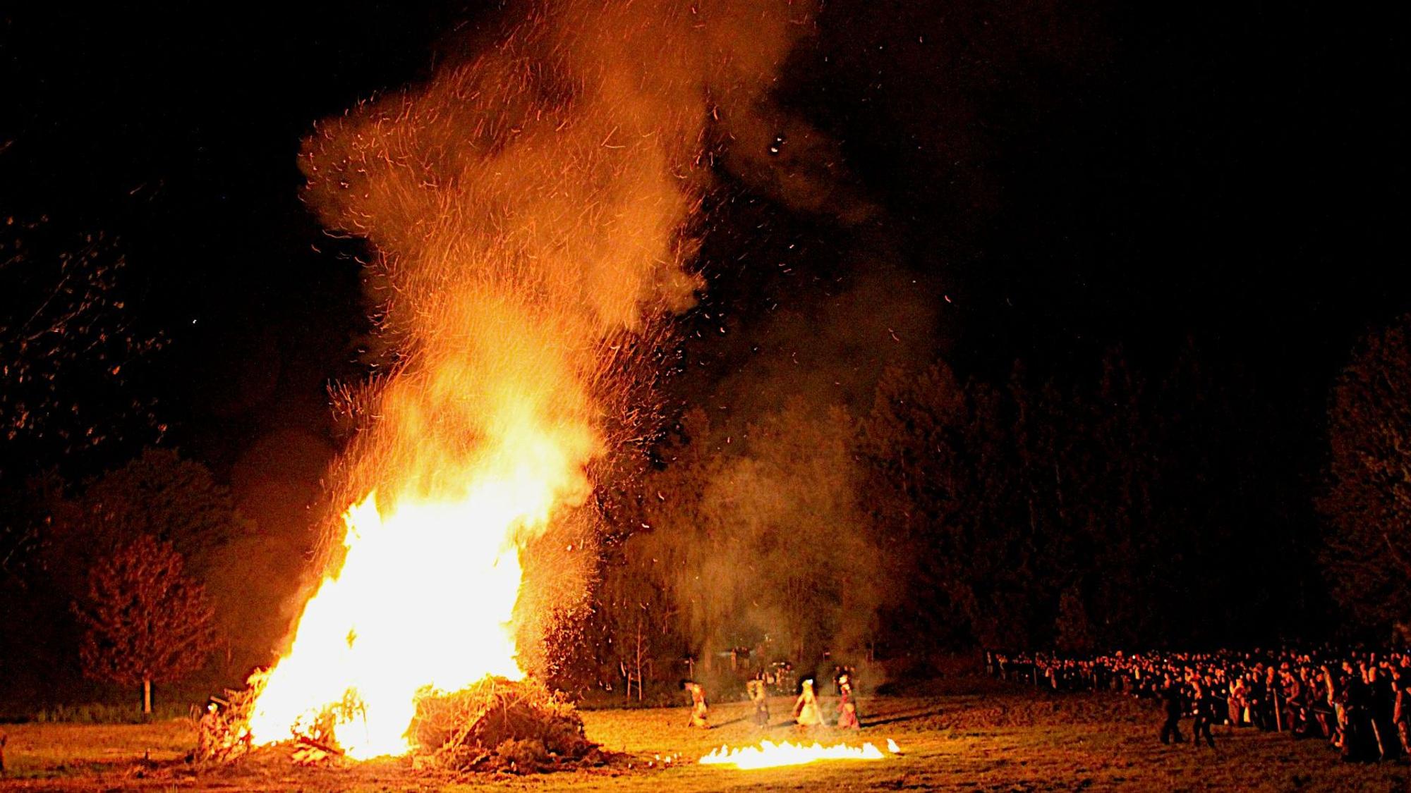 Hoch lodern die Flammen in den nächtlichen Himmel, davor tanzen die vier Hexen. An der Seite verfolgen die Zuschauer das Spektakel.