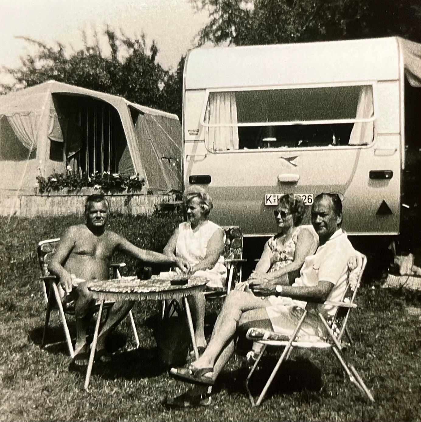 Ein Schwarz-Weiß-Bild zeigt zwei Paar, die auf Campingmöbeln vor einem Wohnwagen sitzen.