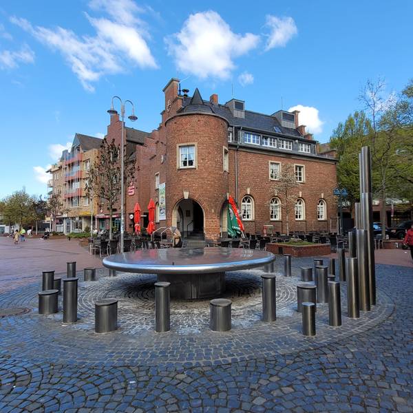 Vorne im Bild ein Brunnen, im Hintergrund ein Backsteingebäude.