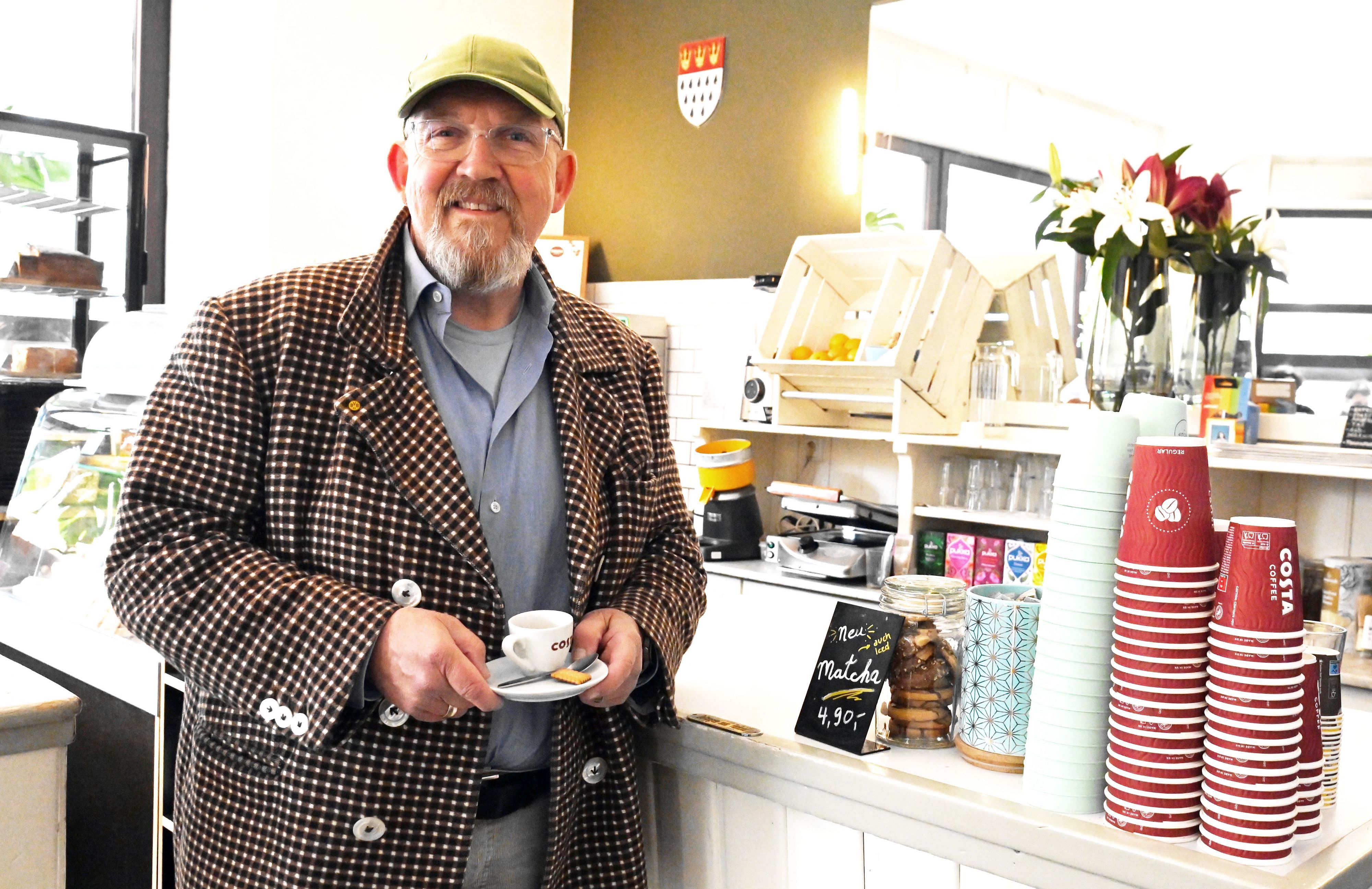 Ein Mann im karierten Sakko steht vor der Theke einer Kaffeebar. In der Hand hält er eine Espresso-Tasse.