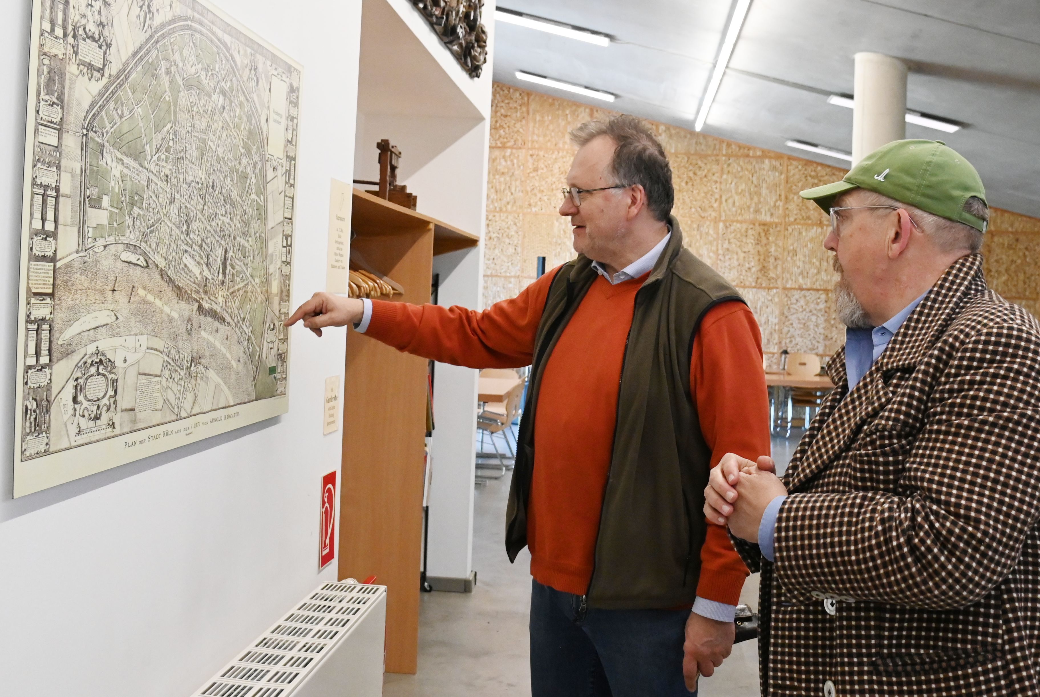 Ein Mann im roten Pullover und Weste zeigt einem Mann mit Bart und Kappe Orte auf einer Karte, die an der Wand hängt.