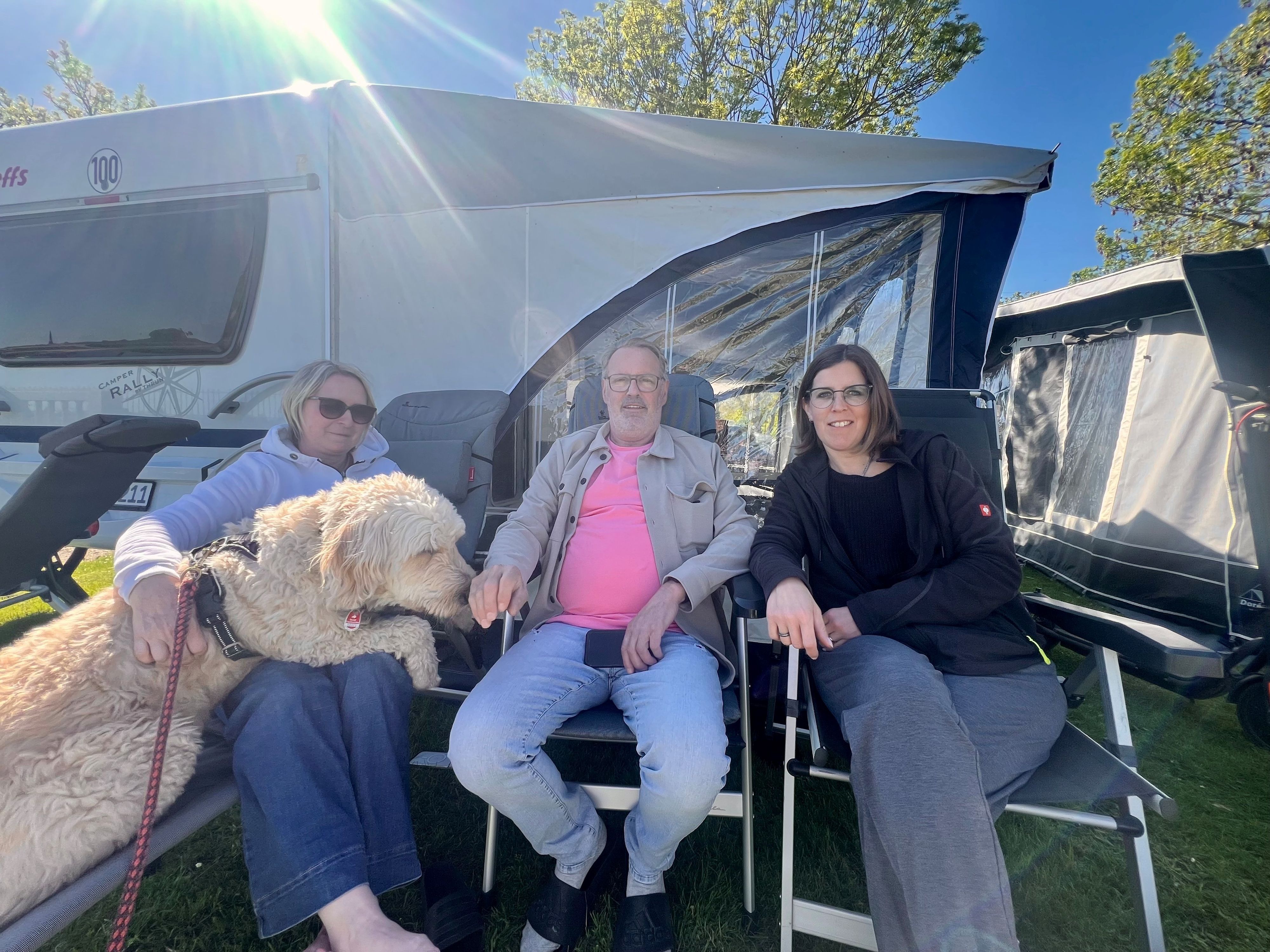 Zwei Frauen, ein Mann und ein großer zotteliger Hund sitzen auf Campingstühlen vor einem Zelt.