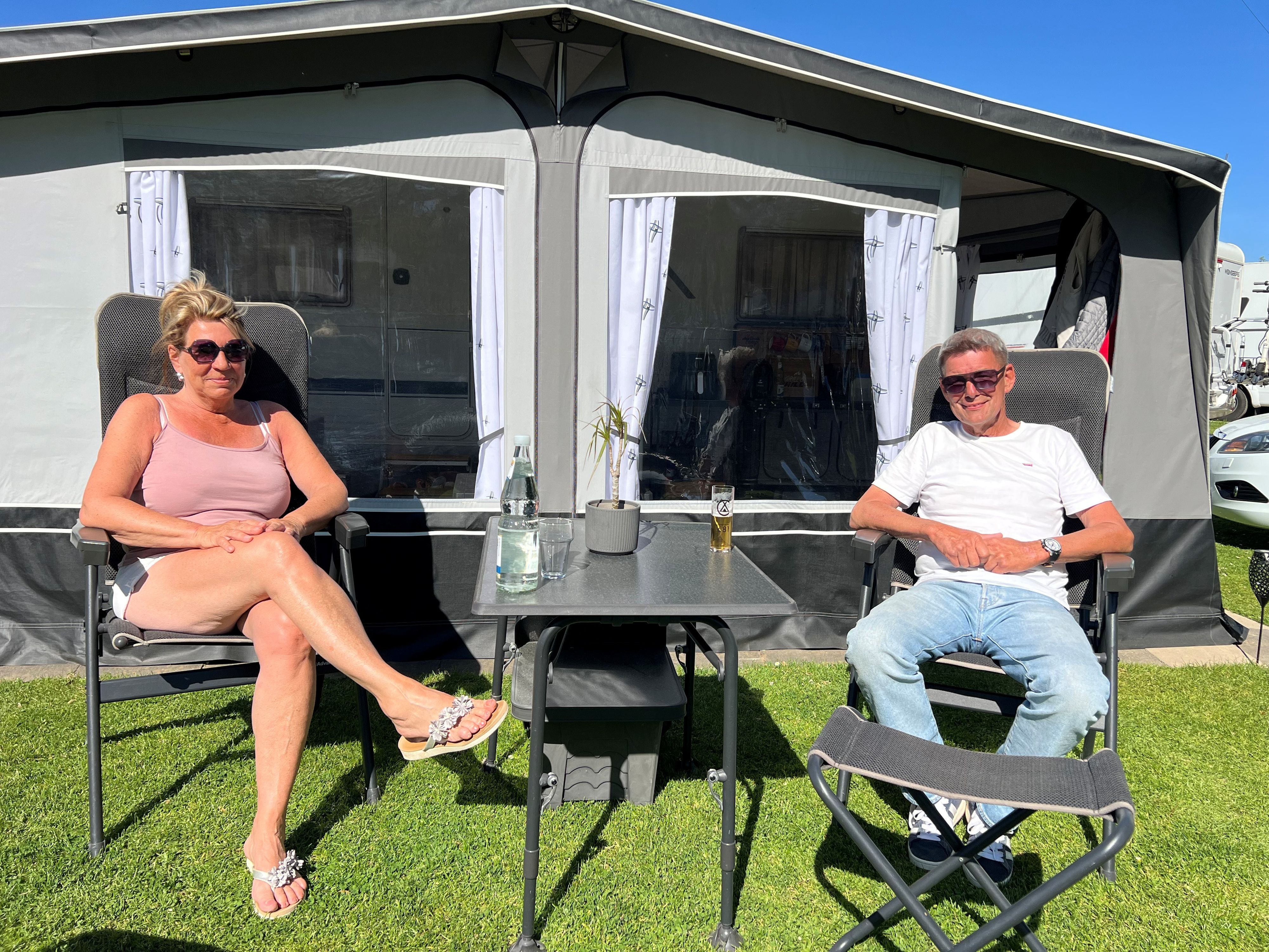 Eine Frau und ein Mann sitzen vor einem Wohnwagen, zwischen sich ein Campingtisch mit Kölsch und Mineralwasser darauf.