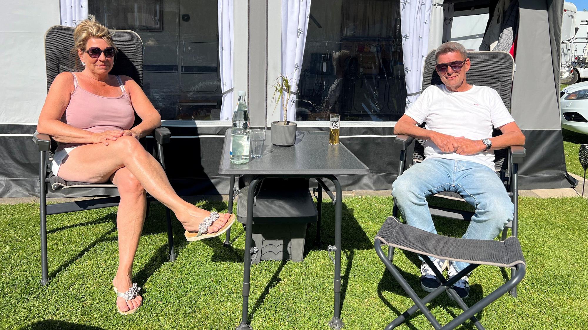 Eine Frau und ein Mann sitzen vor einem Wohnwagen, zwischen sich ein Campingtisch mit Kölsch und Mineralwasser darauf.