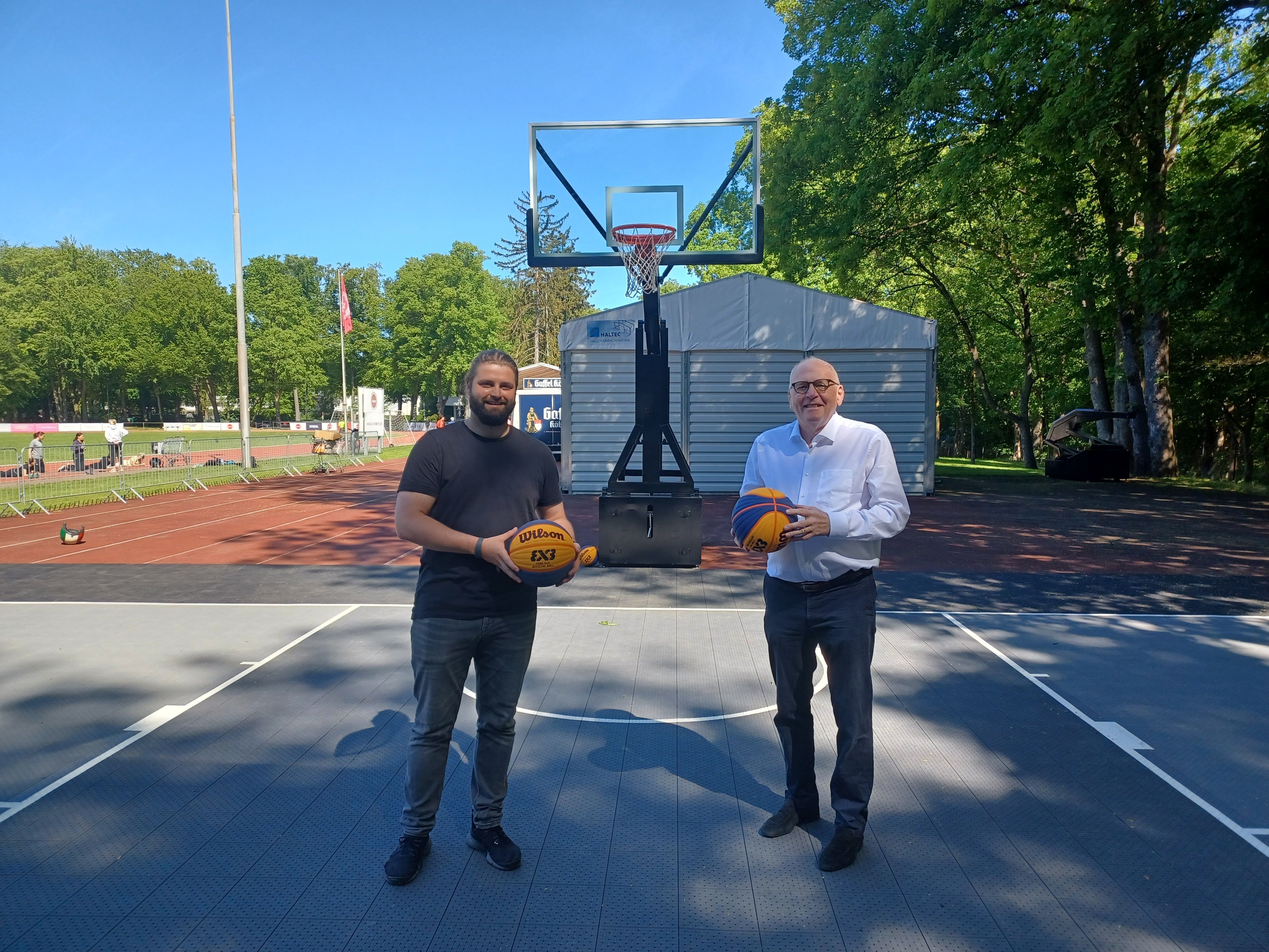Zwei Männer halten Basketbälle in ihren Händen und stehen auf einem neuen 3x3-Basketballplatz.