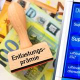 Ein Stempel mit der Aufschrift „Entlastungsprämie“ liegt auf Euroscheinen, daneben steht eine Preistafel an einer Tankstelle.