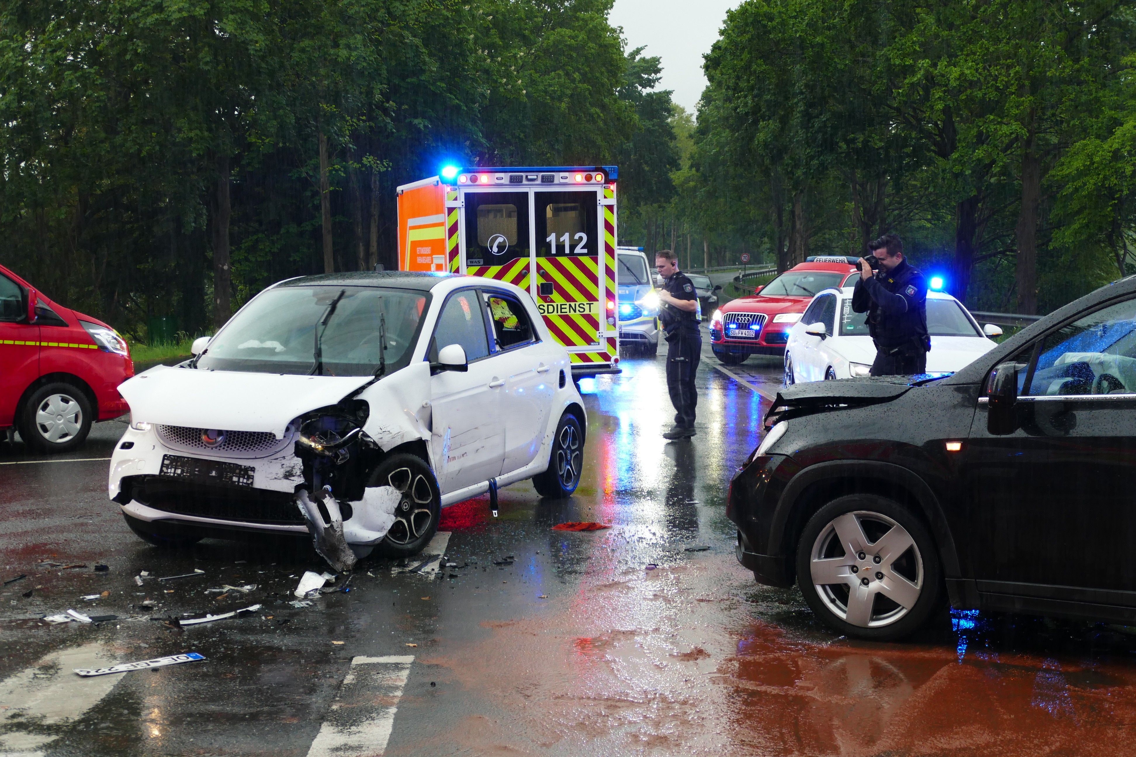 Ein schwarzer und ein weißer Wagen stehen mit zerbeulten Motorhauben auf einer Kreuzung. Um sie herum sind Einsatzkräfte der Polizei und Feuerwehr sowie deren Fahrzeuge.