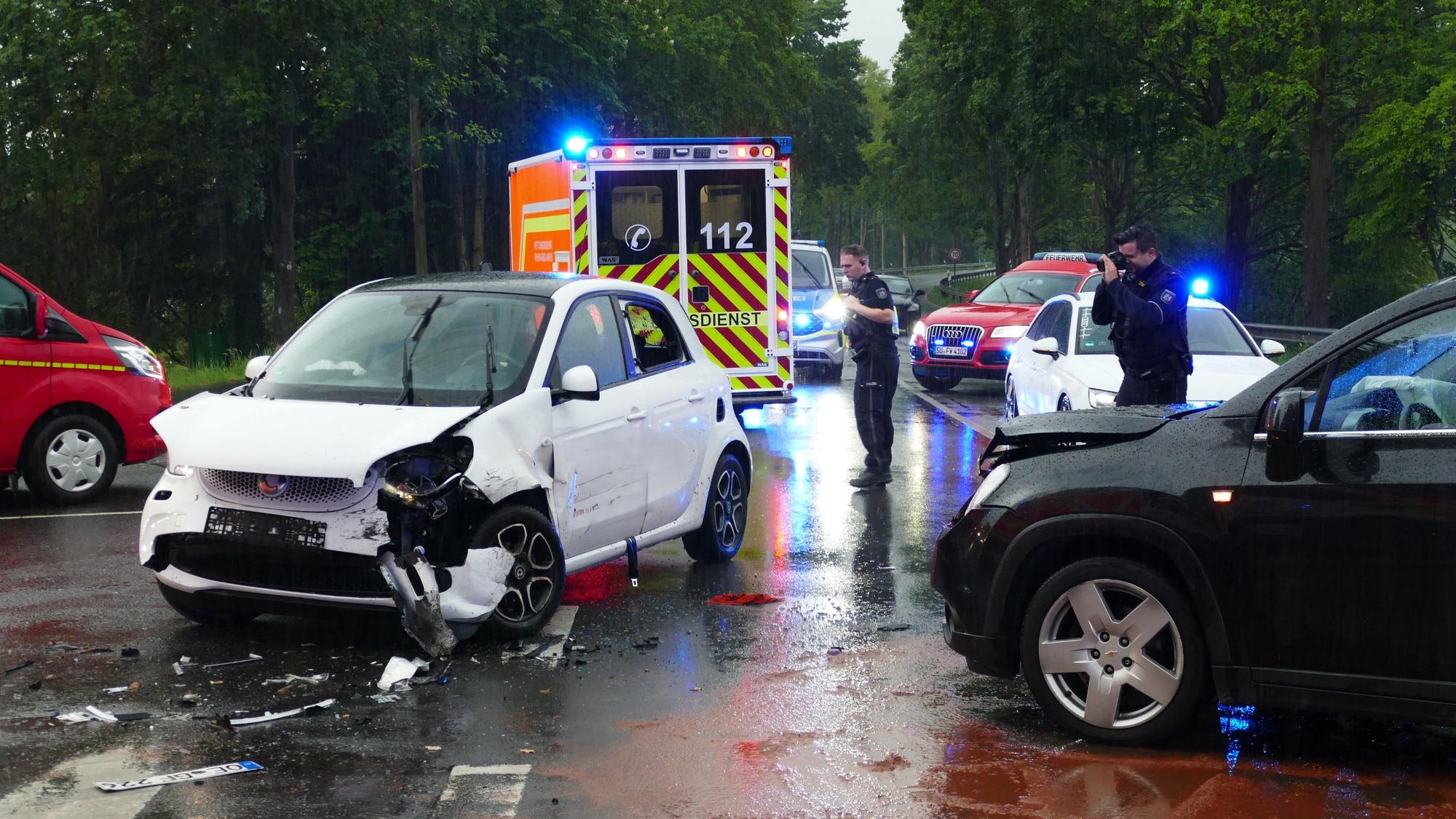 Ein schwarzer und ein weißer Wagen stehen mit zerbeulten Motorhauben auf einer Kreuzung. Um sie herum sind Einsatzkräfte der Polizei und Feuerwehr sowie deren Fahrzeuge.