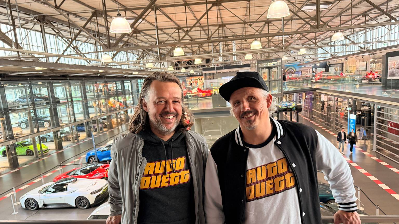 Zwei Männer stehen nebeneinander am Geländer einer Galerie, hinter ihnen sind in einer großen Ausstellungshalle verschiedene Automodelle.