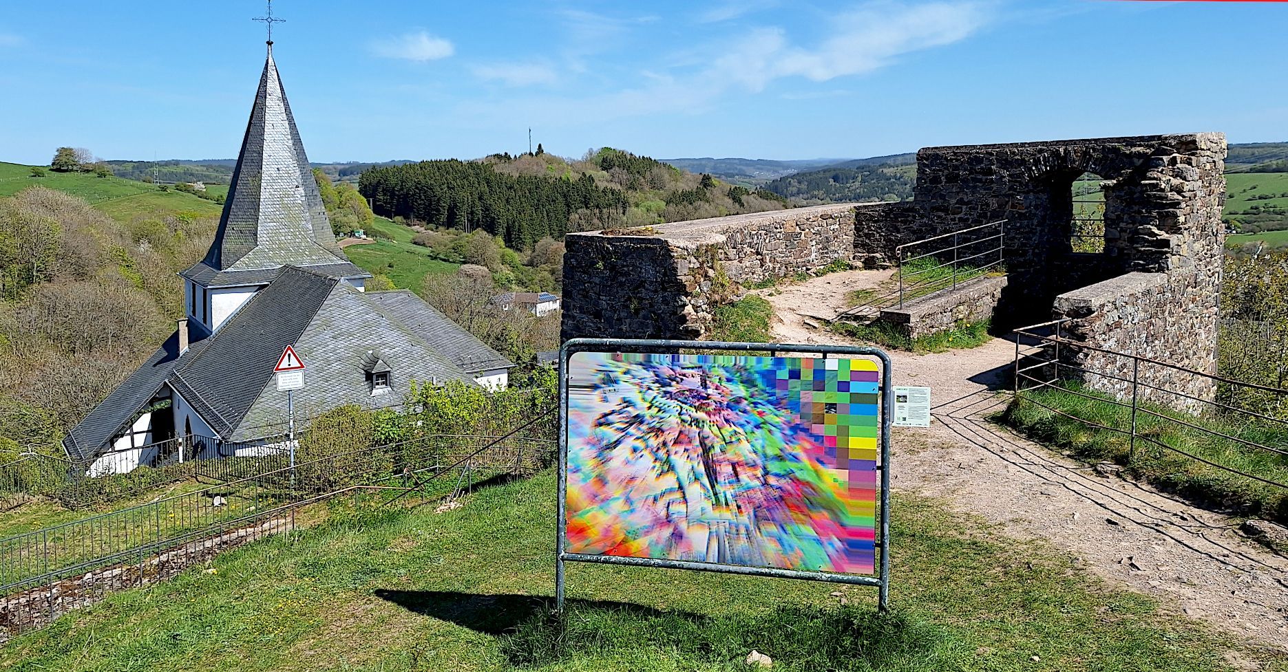 Das Bild zeigt ein Kunstwerk. Im Hintergrund ist die Ruine des Bergfrieds der Kronenburger Burg zu sehen.