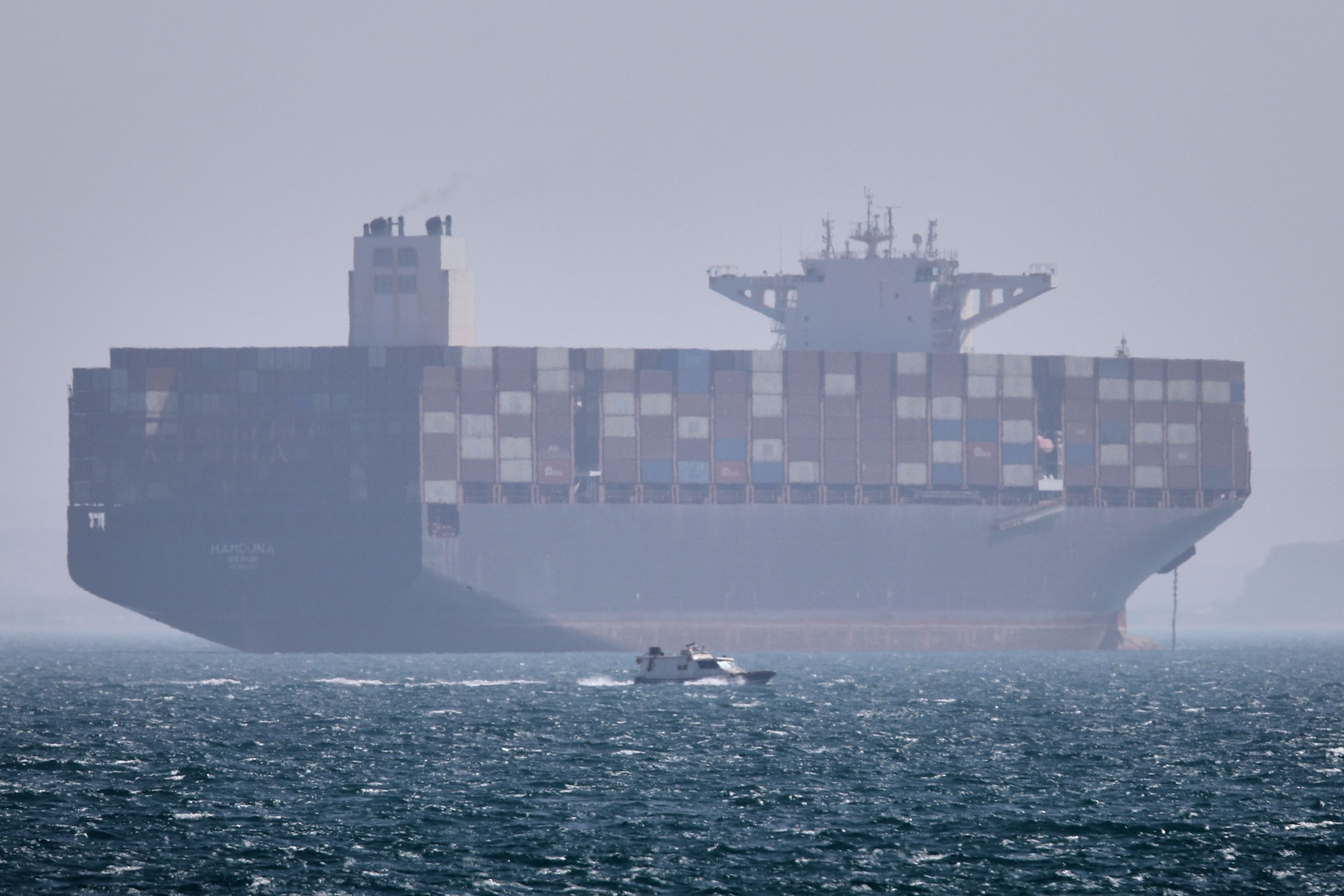 Iran, Bandar Abbas: Ein Containerschiff liegt vor Anker, während im Vordergrund ein kleines Motorboot in der Straße von Hormus vorbeifährt (Archivbild).