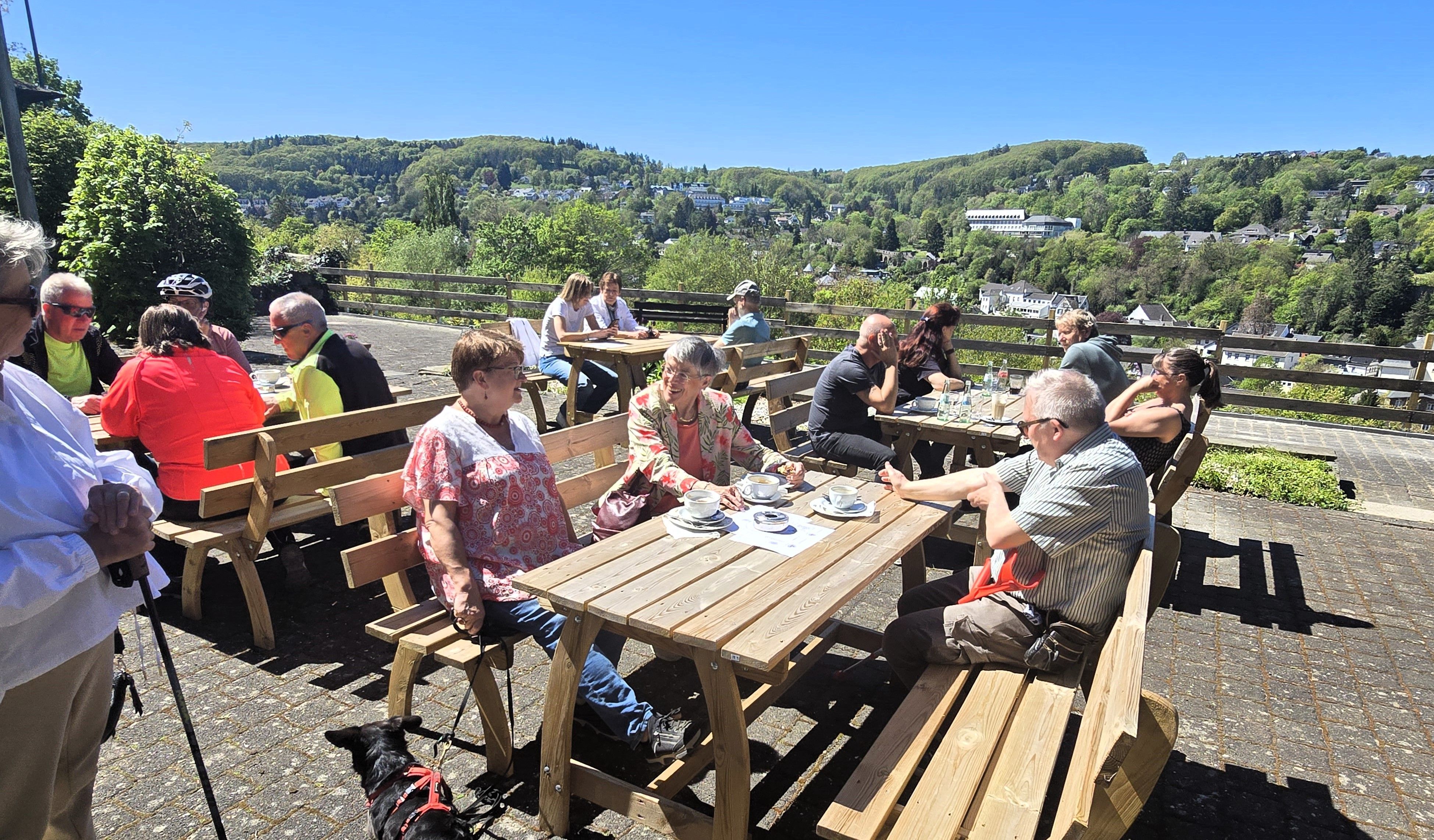Menschen sitzen auf einer Café-Terrasse in der Sonne und genießen Kaffee, Kuchen und den Ausblick auf Bad Münstereifel.