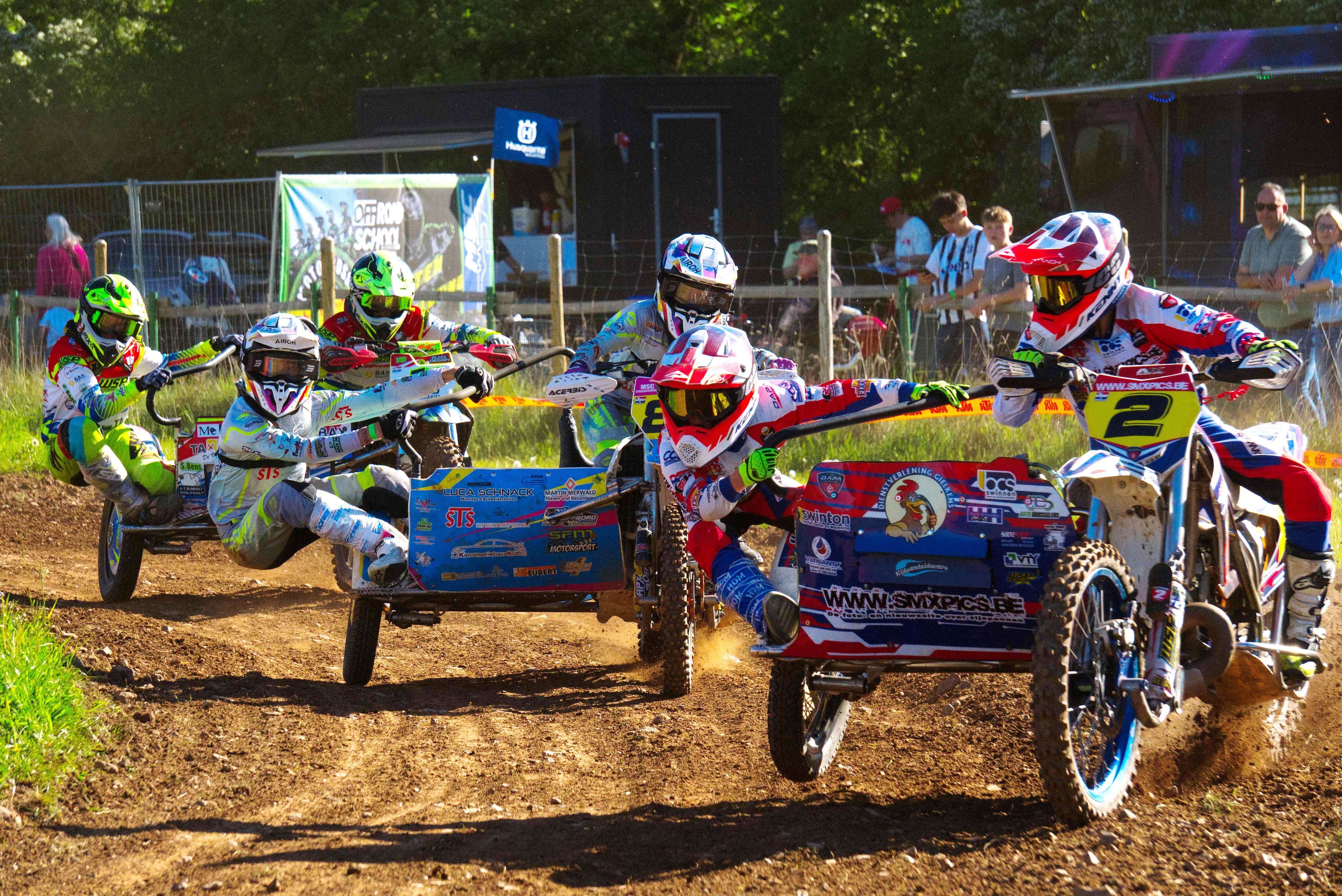 Drei Motocross-Maschinen mit Seitenwagen fahren durch eine Kurve, die Beifahrer hängen artistisch am Seitenwagen.