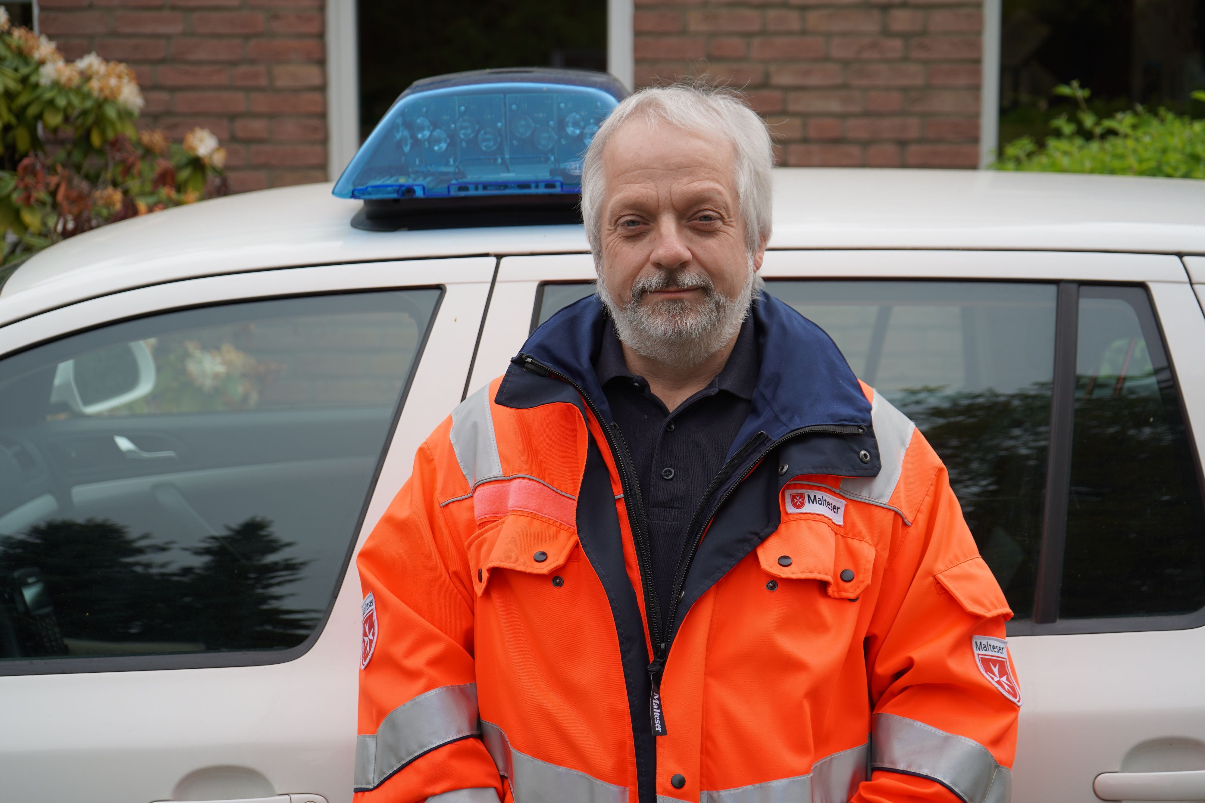 Das Bild zeigt Hornfeck mit einer orangen Jacke vor einem Dienstwagen mit Blaulicht.