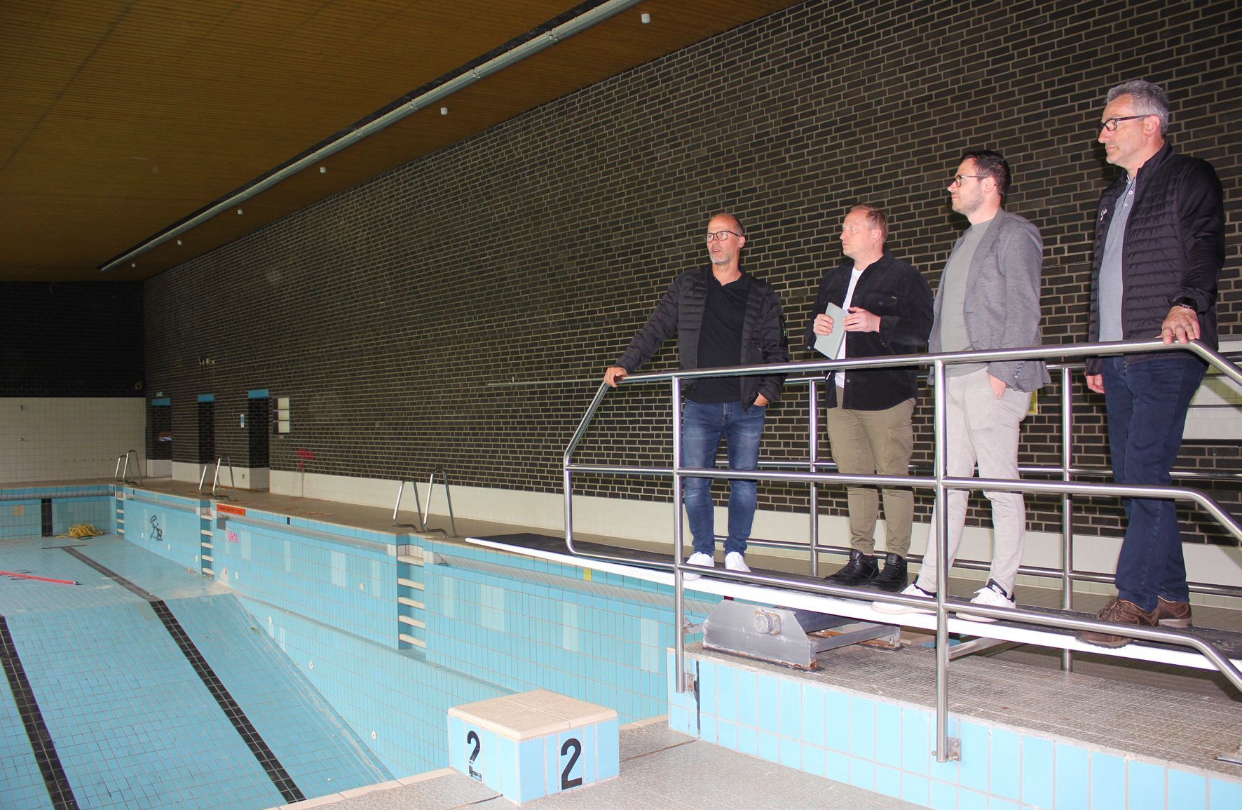 Bei einem Ortstermin im Schwimmbad stehen die vier genannten Männer auf dem Sprungbrett über dem leeren Becken.