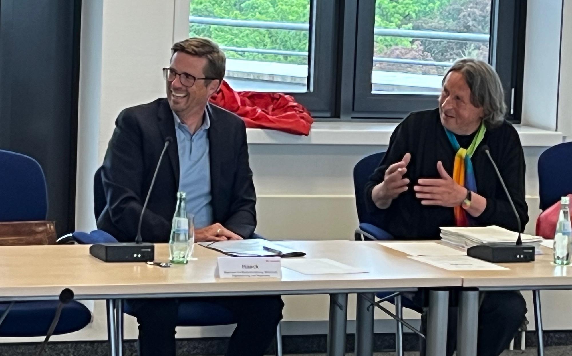 Ein Mann mit Brille und dunklem Jackett sitzt neben einem Mann mit kinnlangem Haar, schwarzem Pullover und buntem Schal.