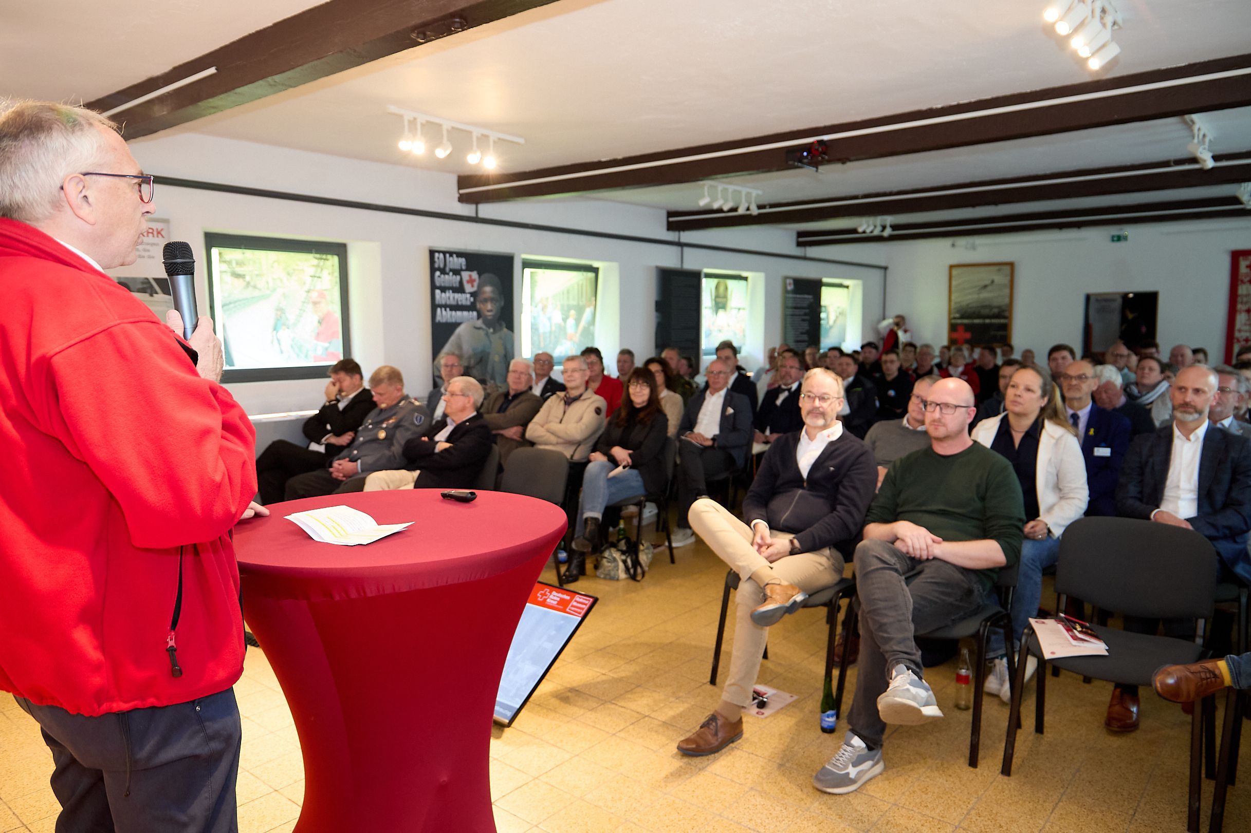 Karl-Werner Zimmermann steht an einem Tisch mit roter Husse und spricht im Rotkreuzmuseum vor einem vollen Auditorium.