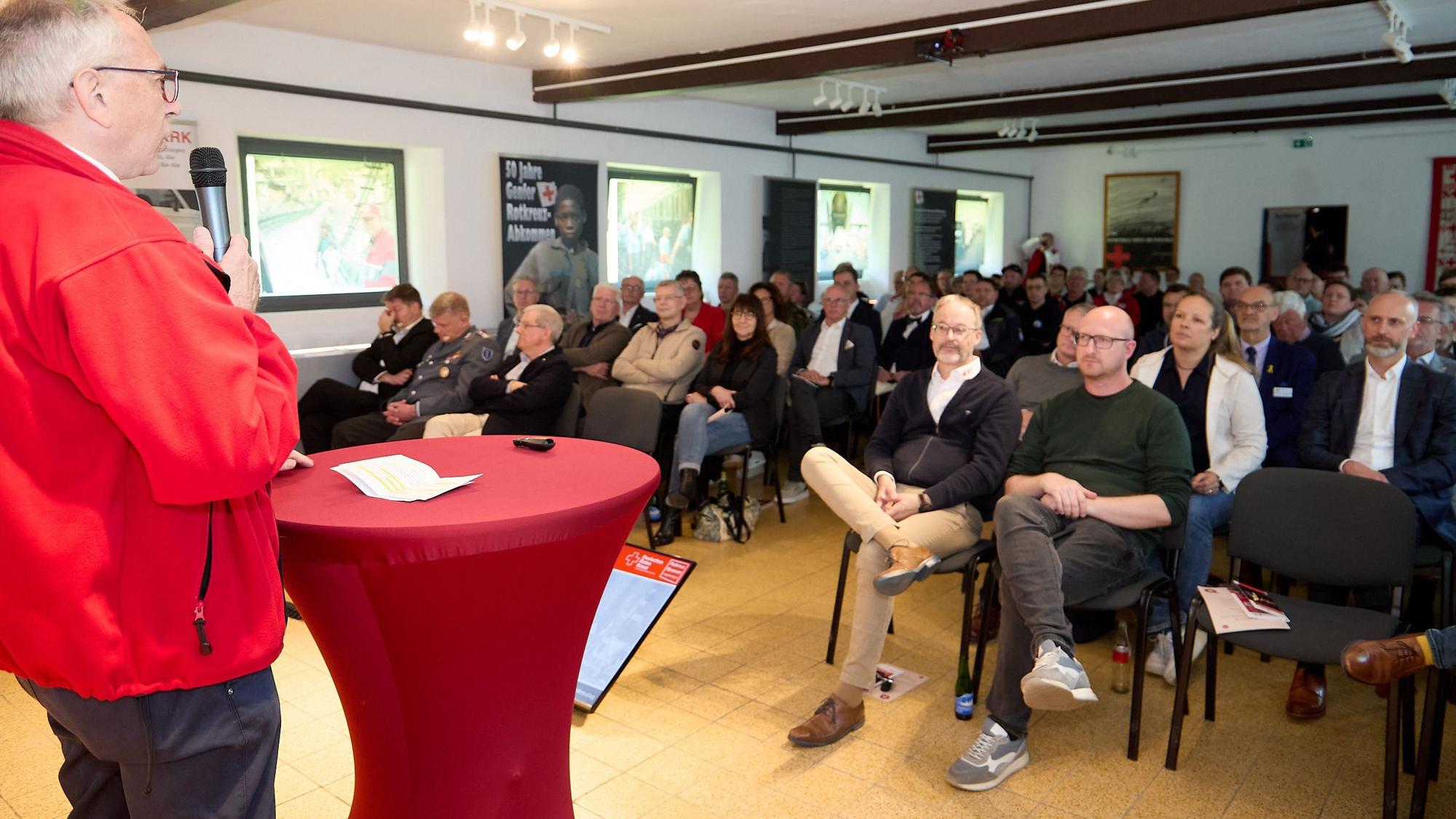 Karl-Werner Zimmermann steht an einem Tisch mit roter Husse und spricht im Rotkreuzmuseum vor einem vollen Auditorium.