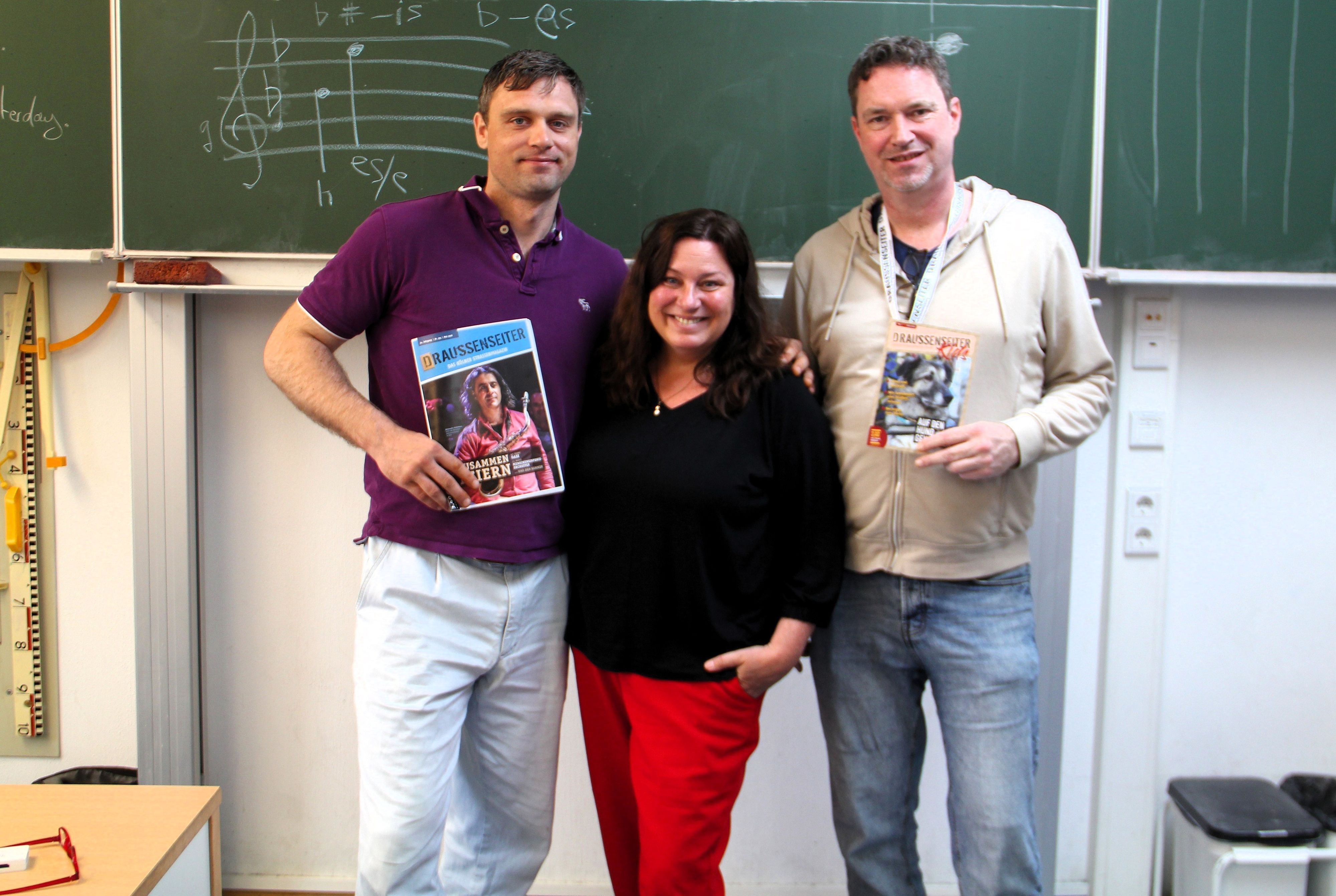Zwei Männer und eine Frau stehen vor einer Tafel in einem Klassenzimmer.