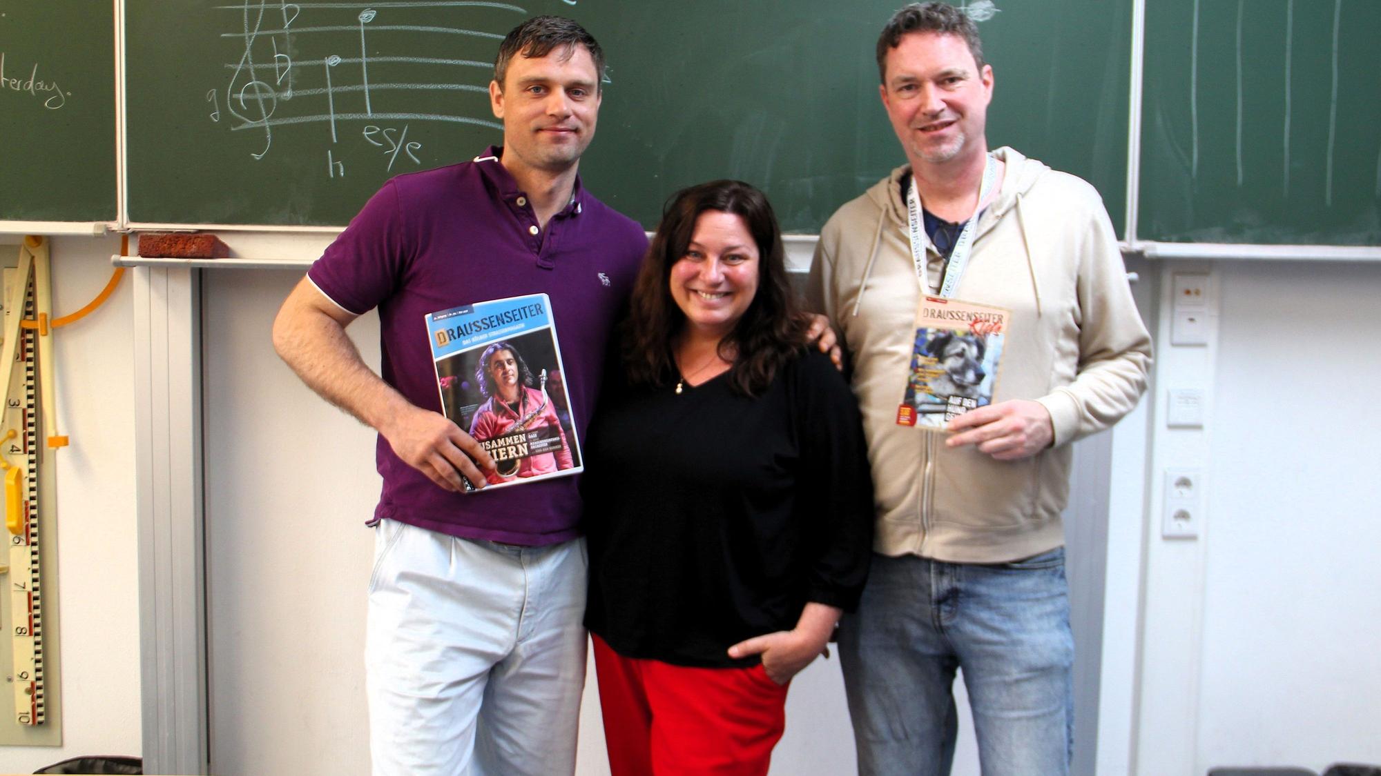 Zwei Männer und eine Frau stehen vor einer Tafel in einem Klassenzimmer.