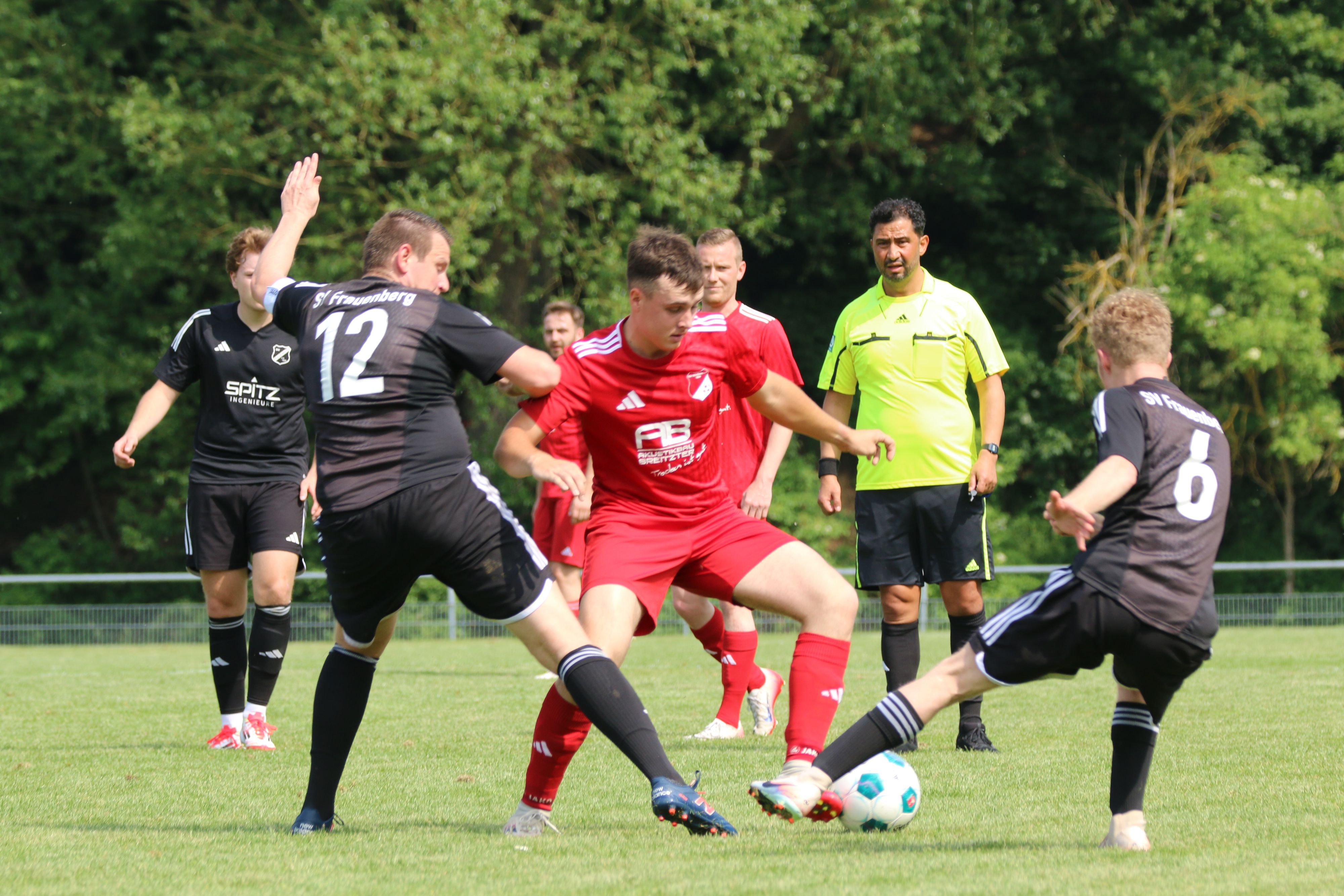 Fußballer Tim Breuer vom TSV Schönau im roten Trikot setzt sich  gegen zwei Spieler des SV Frauenberg in schwarzen Trikots durch.