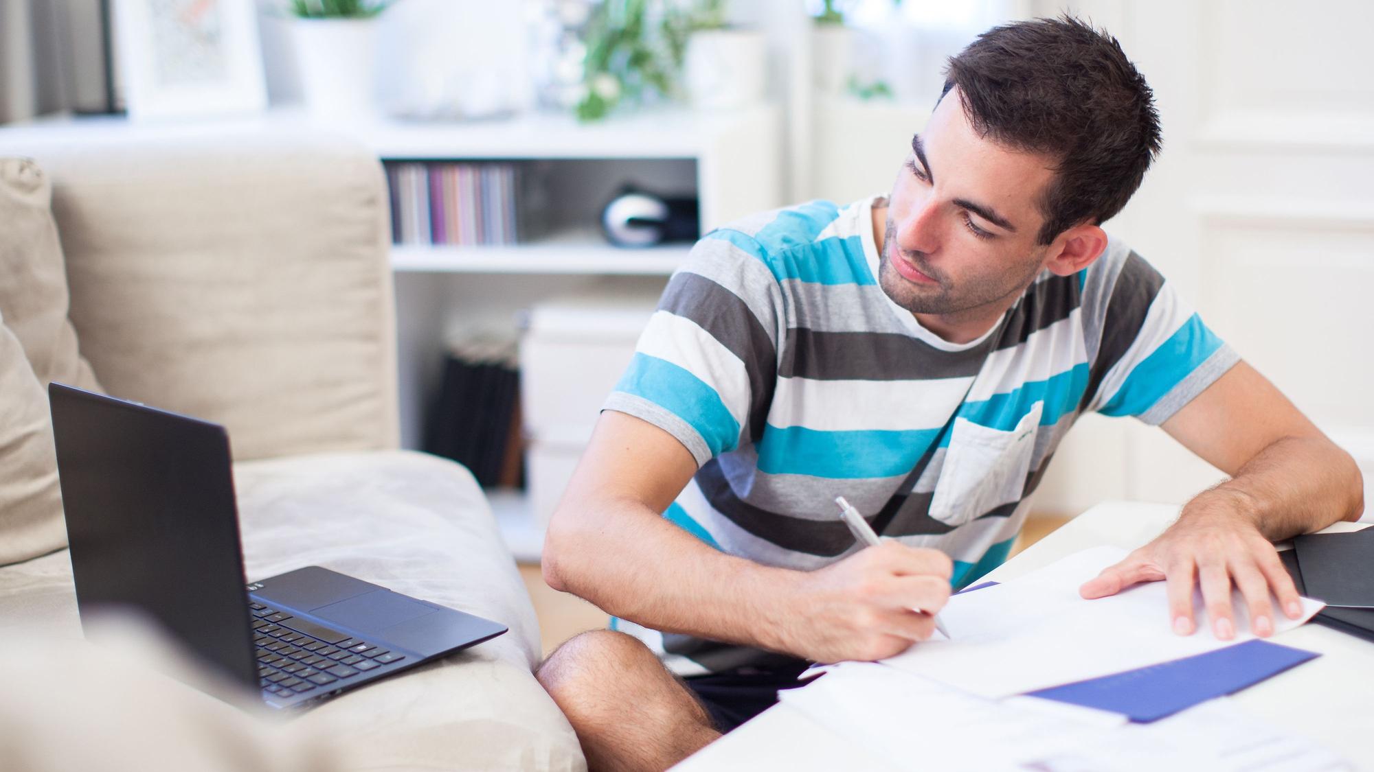 Ein Mann sitzt vor einem Laptop, der auf einem Sofa steht, und schreibt auf einem Papier.