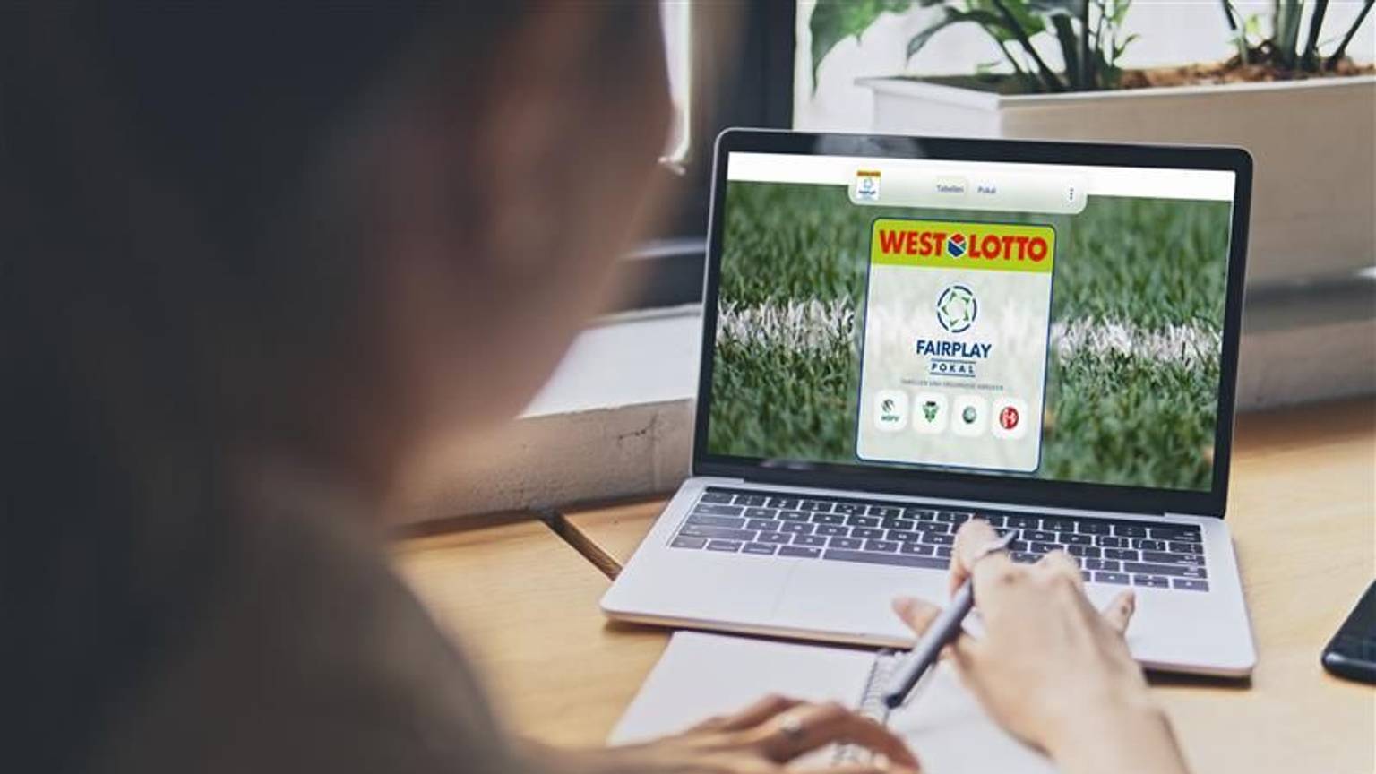 Eine Frau sitzt an einem Tisch und schaut auf ihren Laptop. Der Laptop zeigt die Internetseite des WestLotto Fairplay-Pokals.
