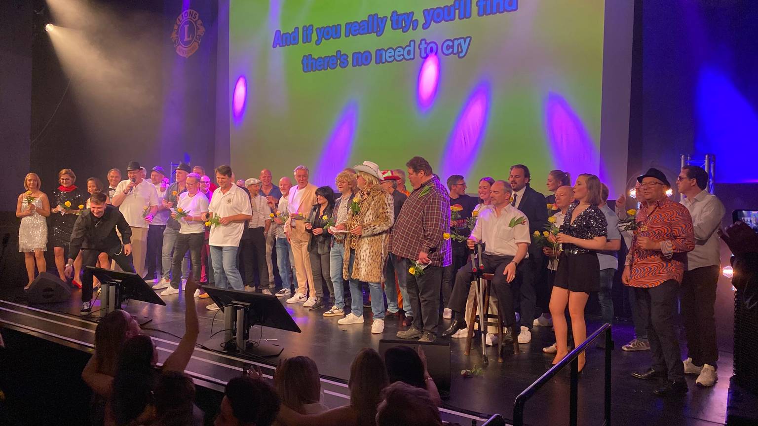 etwa 30 Menschen auf einer erleuchteten Bühne, im Hintergrund eine große Leinwand