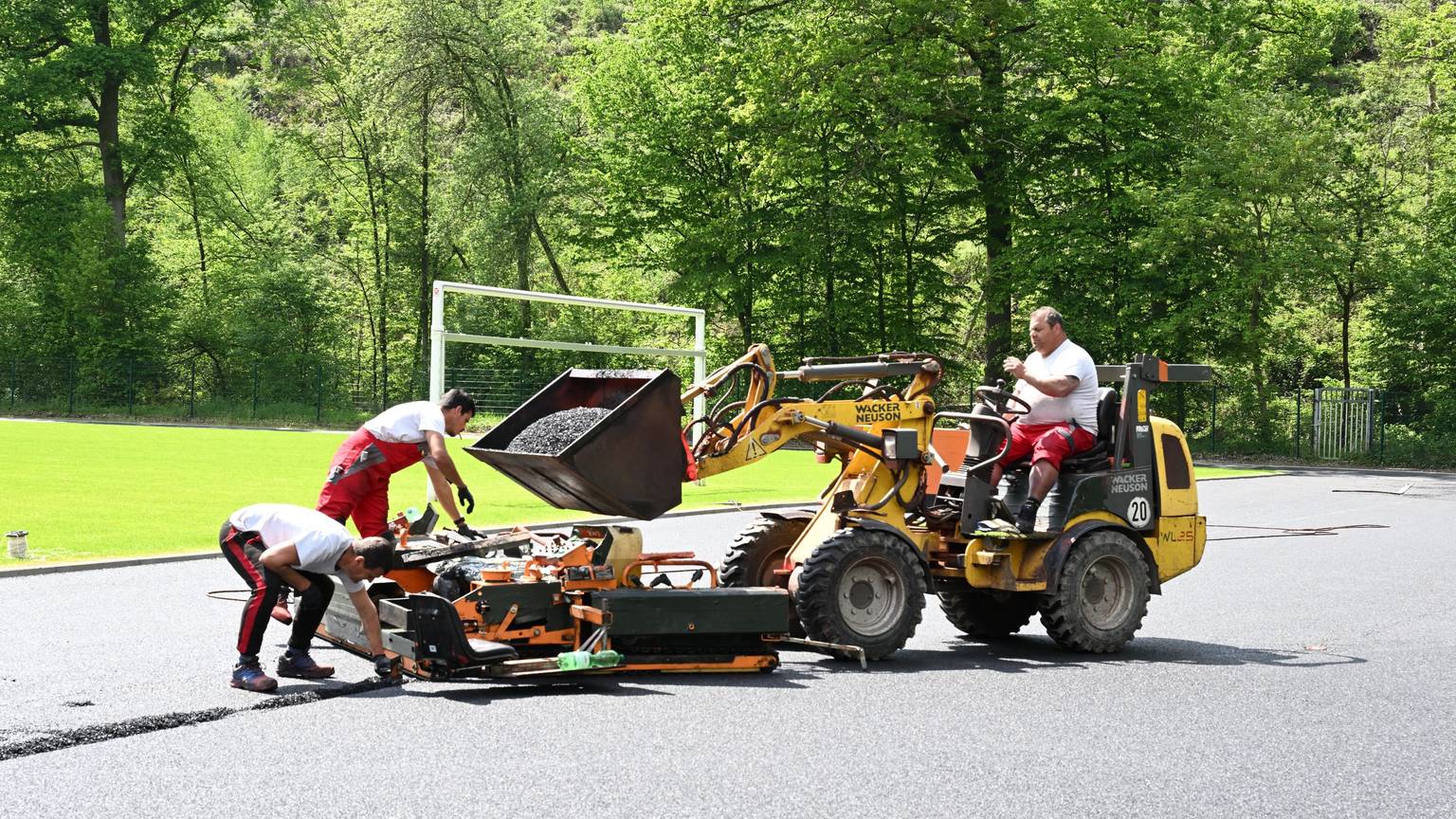 Ein Mann sitzt auf einem Schaufelbagger und lädt dunkles Granulat in eine Mschine, mit der zwei andere Männer das Material auf den Laufbahnen eines Sportplatzes verteilen.
