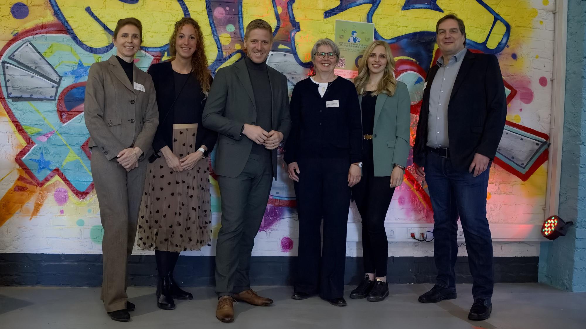 Vier Frauen und zwei Männer stehen in der Zikkurat in Firmenich vor einer Wand, auf der ein Graffiti zu sehen ist.