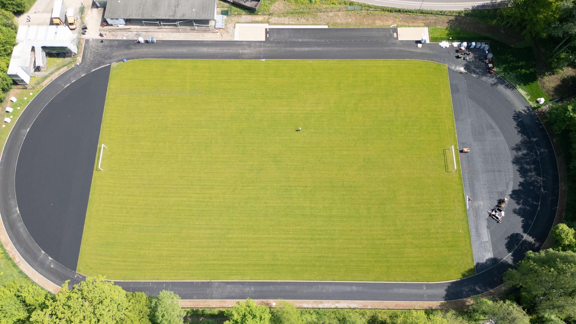 Ein im Bau befindlicher Sportplatz aus der Vorgelperspektive. In der Mitte liegt schon grüner Rollrasen, die Laufbahnen werden gerade mit ein einer grau-schwarzen Kunststoffschicht belegt.