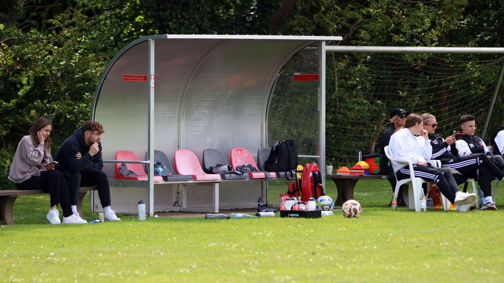 Die leere Ersatzbank der Heimmannschaft. Daneben sitzen Zuschauer, darunter auch verletzte Spielerinnen, auf Stühlen.