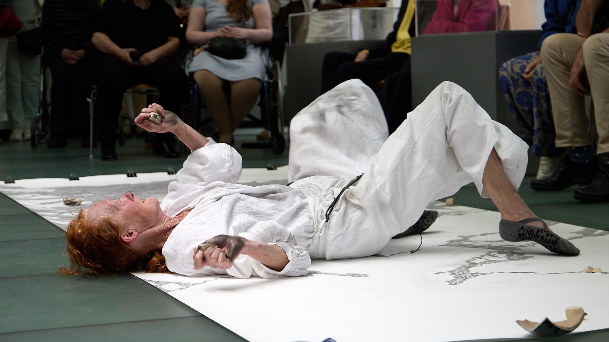 Die Künstlerin, die einen weißen Judo-Anzug trägt, liegt auf dem Rücken und rutscht über eine auf dem Boden ausgerollte Leinwand.