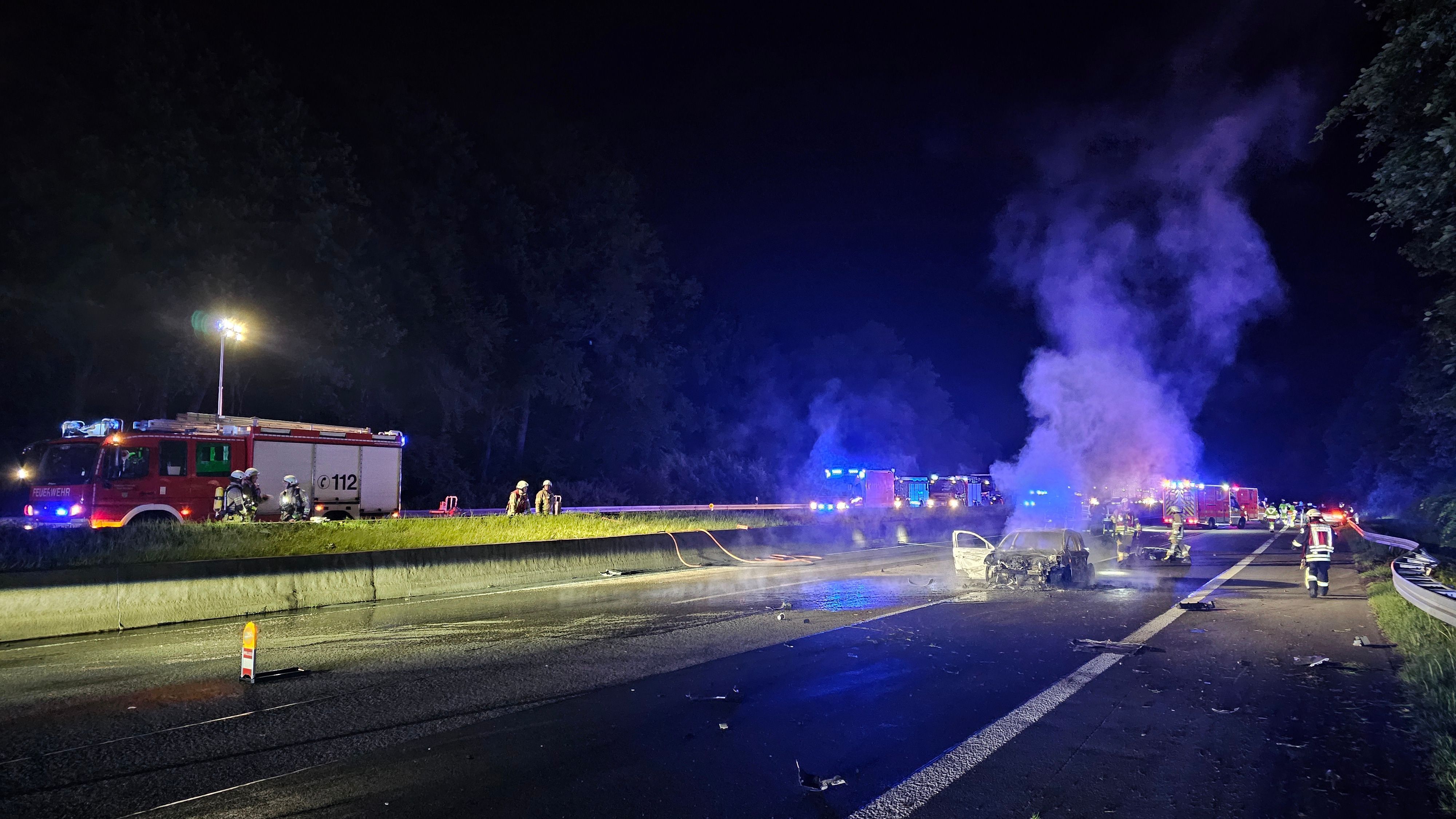 Feuerwehrautos stehen auf einer Autobahn in Höhe eines ausgebrannten Fahrzeugs.